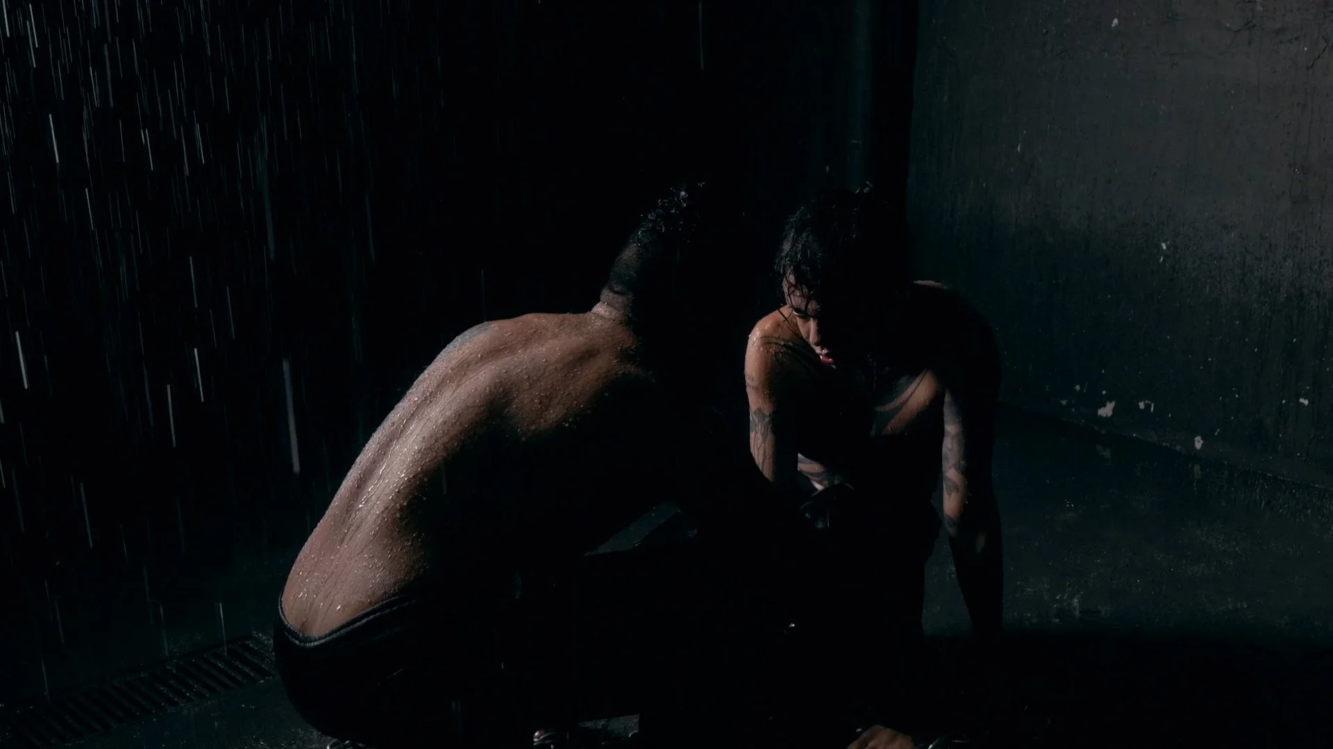 A man and woman sitting on the ground in the rain, with water droplets visible, in a dark setting with rain pouring down.