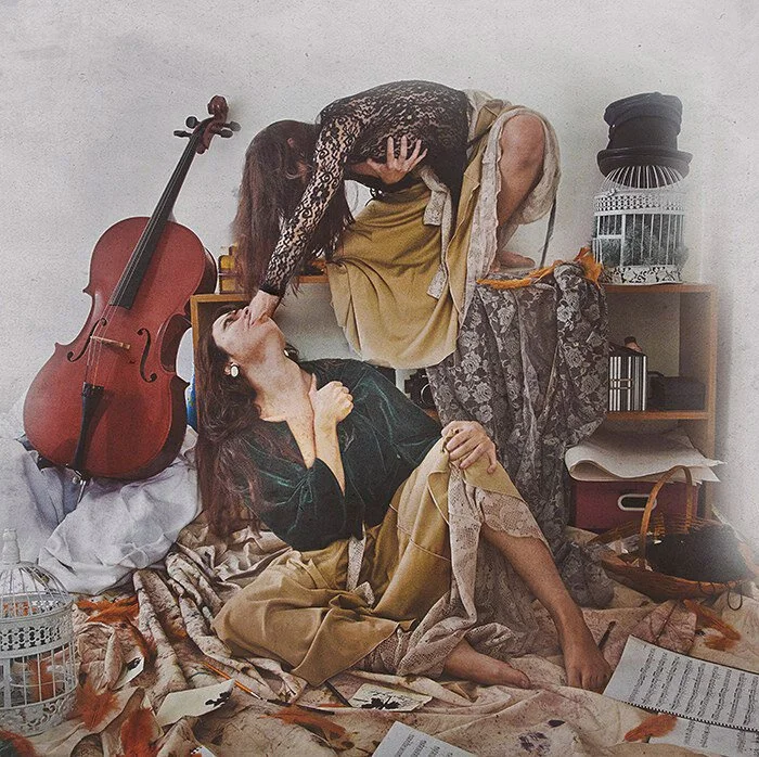 Two women in vintage-style clothing engaging in a playful or dramatic scene in a cluttered room, with a cello leaning against the wall, and various papers, a birdcage, and a basket around them.