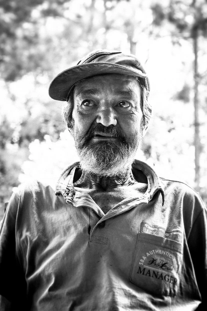 Black and white photo of an older man with a beard, wearing a clubhouse cap and a collared shirt with a patch that says 'Manager,' outdoors with trees in the background.