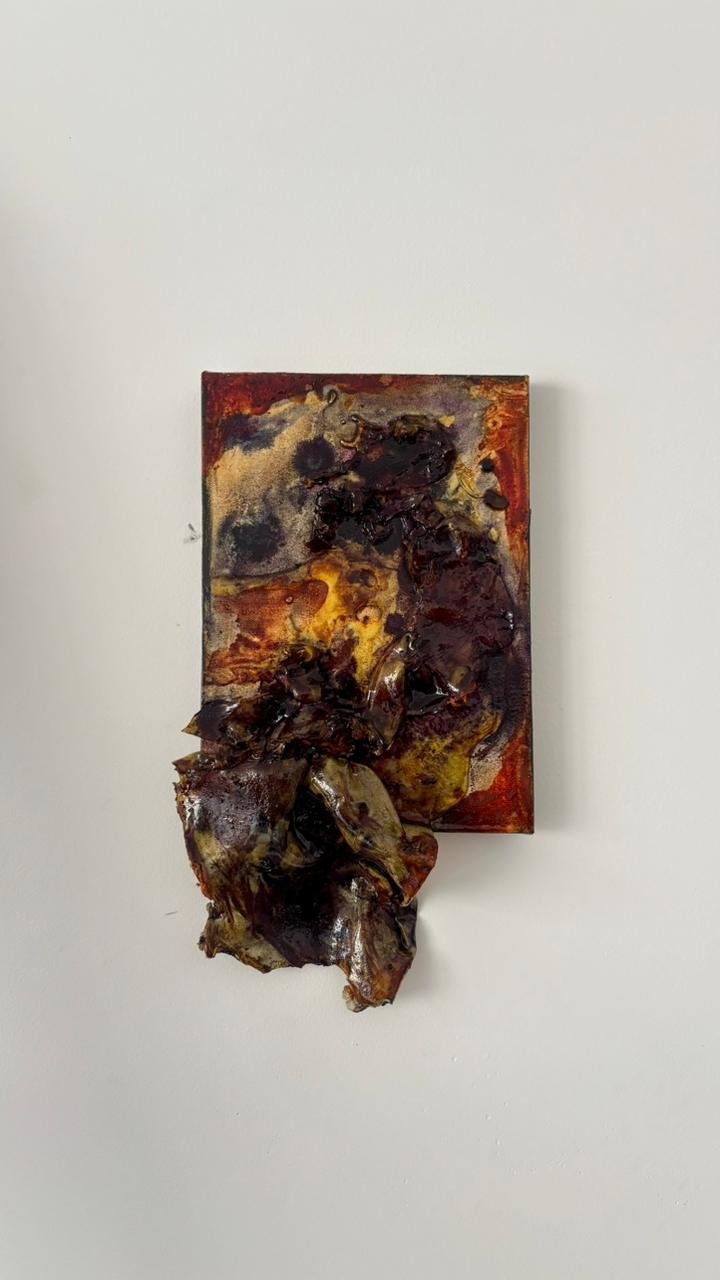 Mixed media artwork featuring a rectangular canvas with abstract dark and warm-colored patterns, with melted or crumpled material hanging from the bottom.