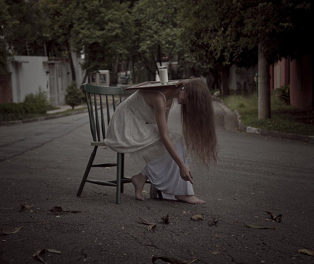 A woman is sitting on a chair on a deserted street, hunched over and touching her foot, with a tray balanced on her head delicately. She has long hair and is wearing a white dress; the scene appears somber and quiet.