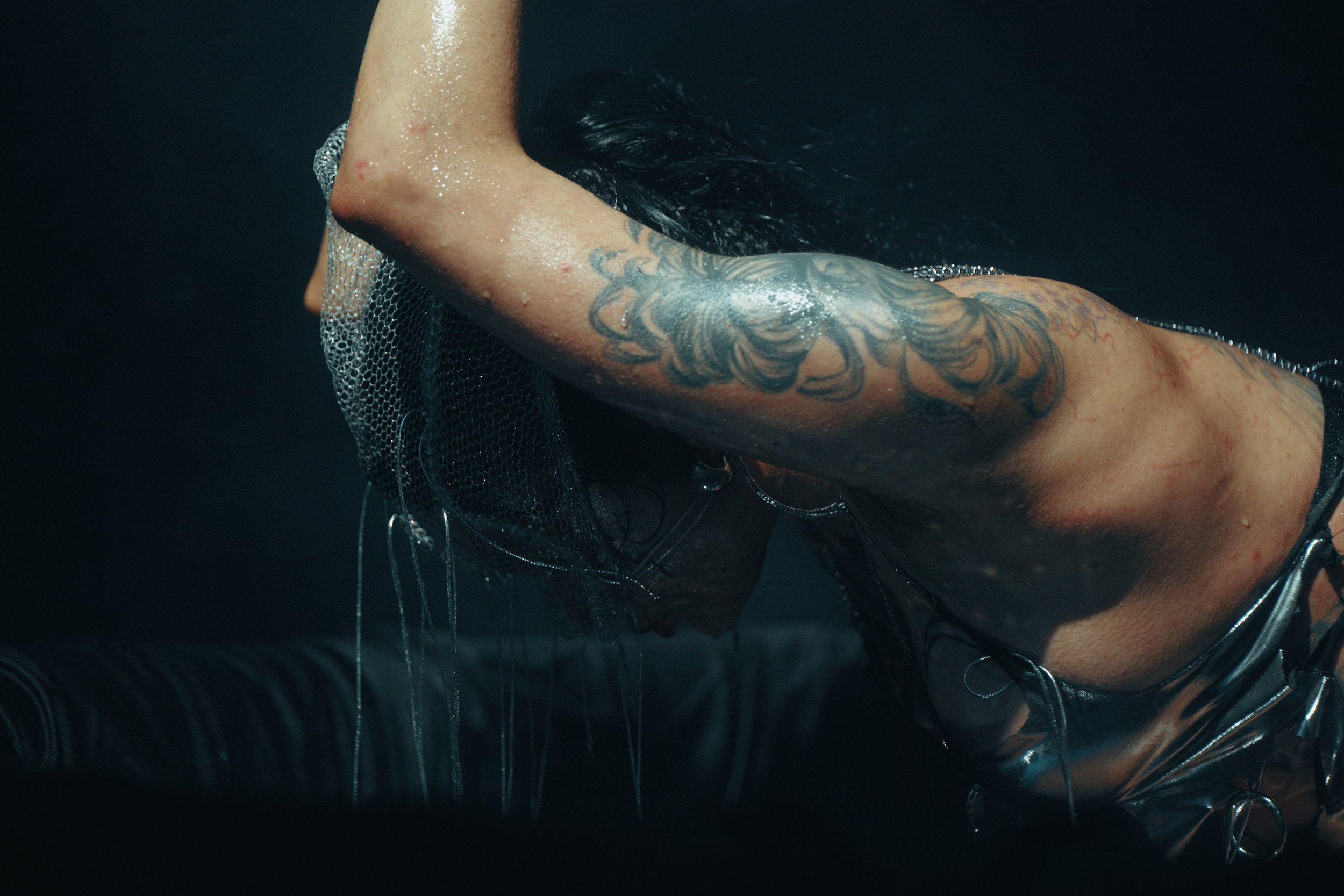 A person with tattoos on their arm leaning forward, wearing wet clothing, with water droplets on their skin and hair.