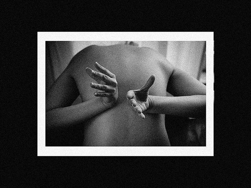 Black and white photo of a nude person from behind, with hands on their back. The image is framed within a white border on a black background.