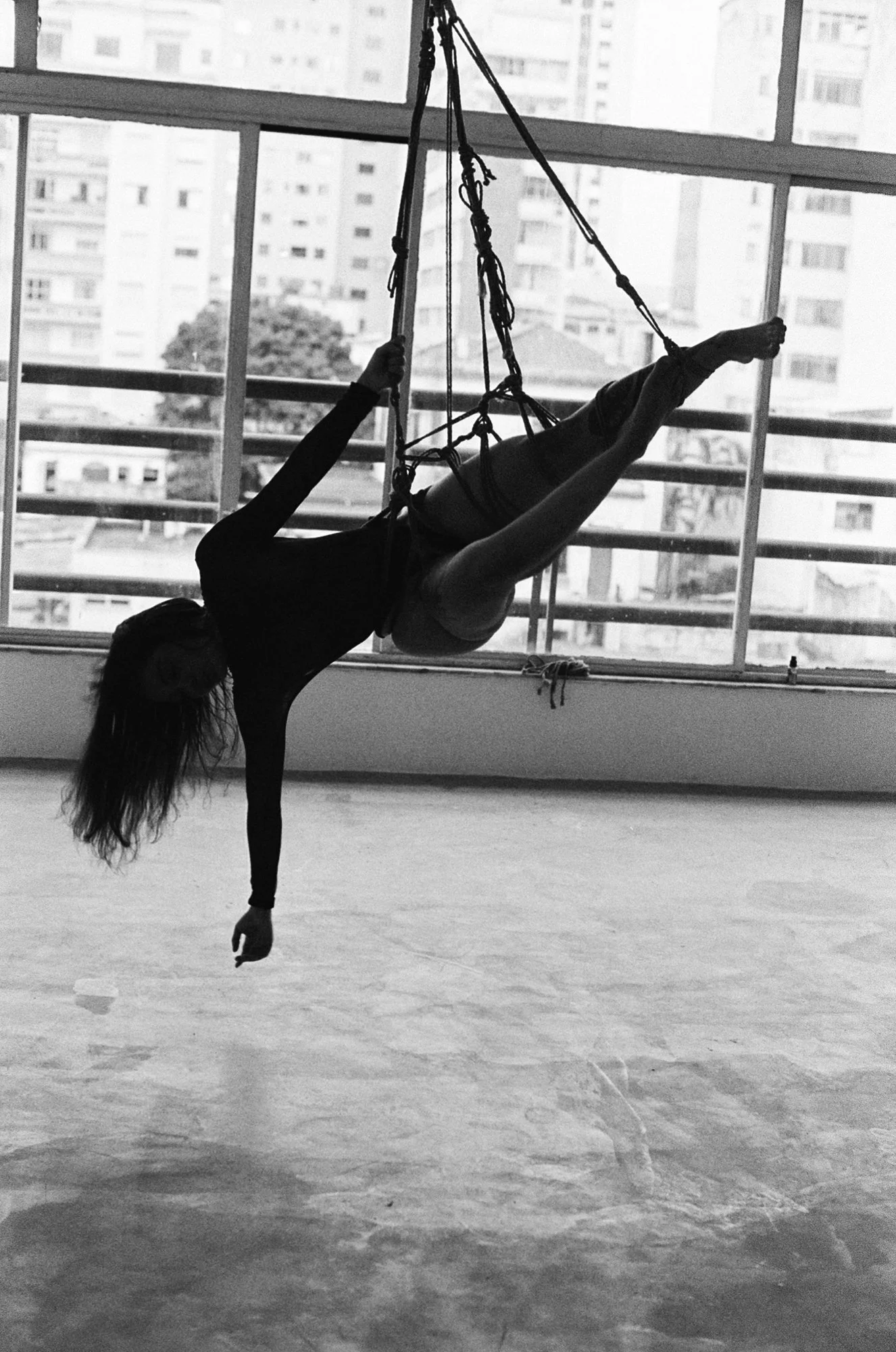 A person in athletic wear practicing aerial silks inside a gym with large windows exposing a cityscape of high-rise buildings.