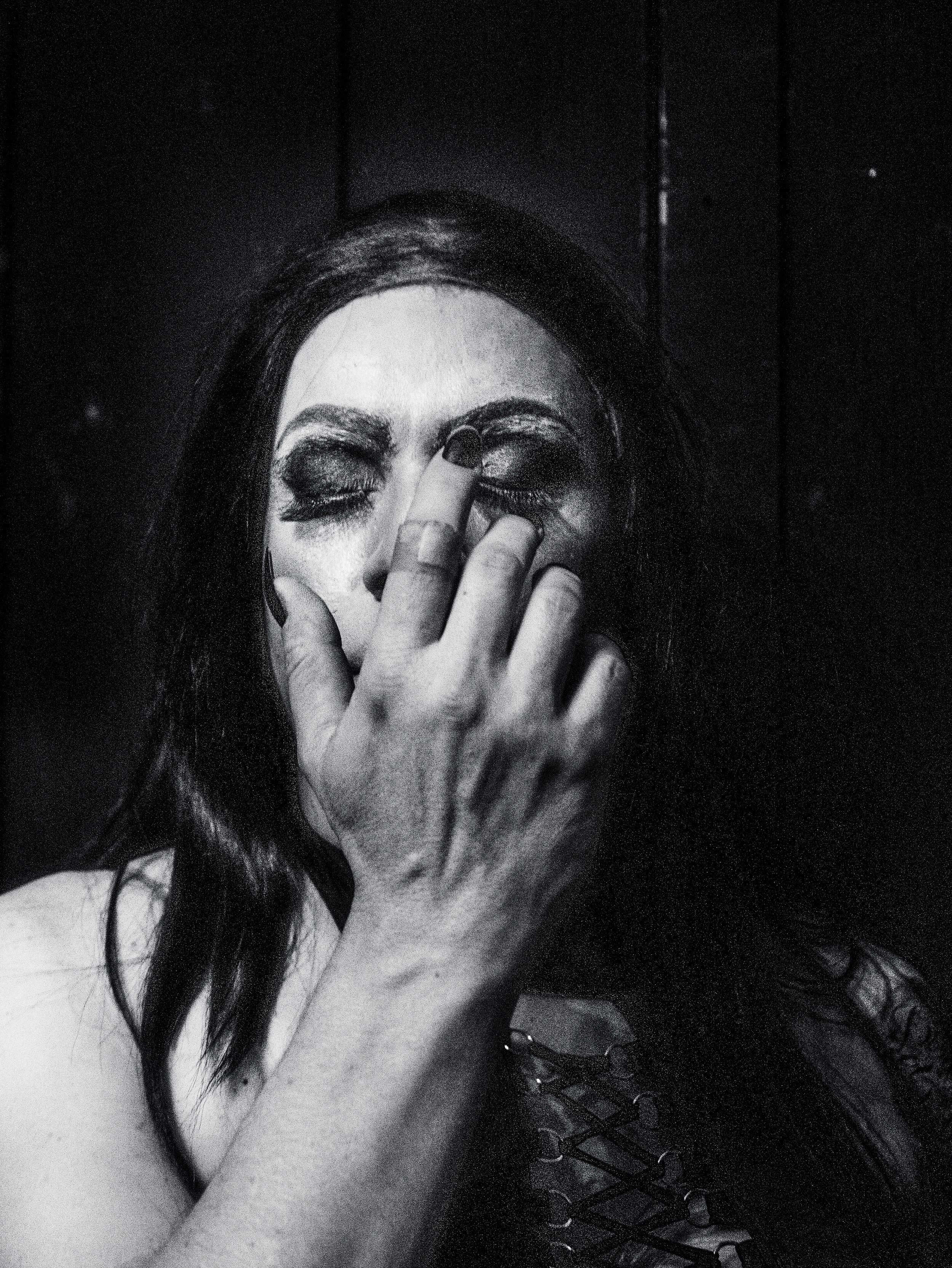 A woman with dark hair and makeup covering her face with her hand, eyes closed, in a black and white image.