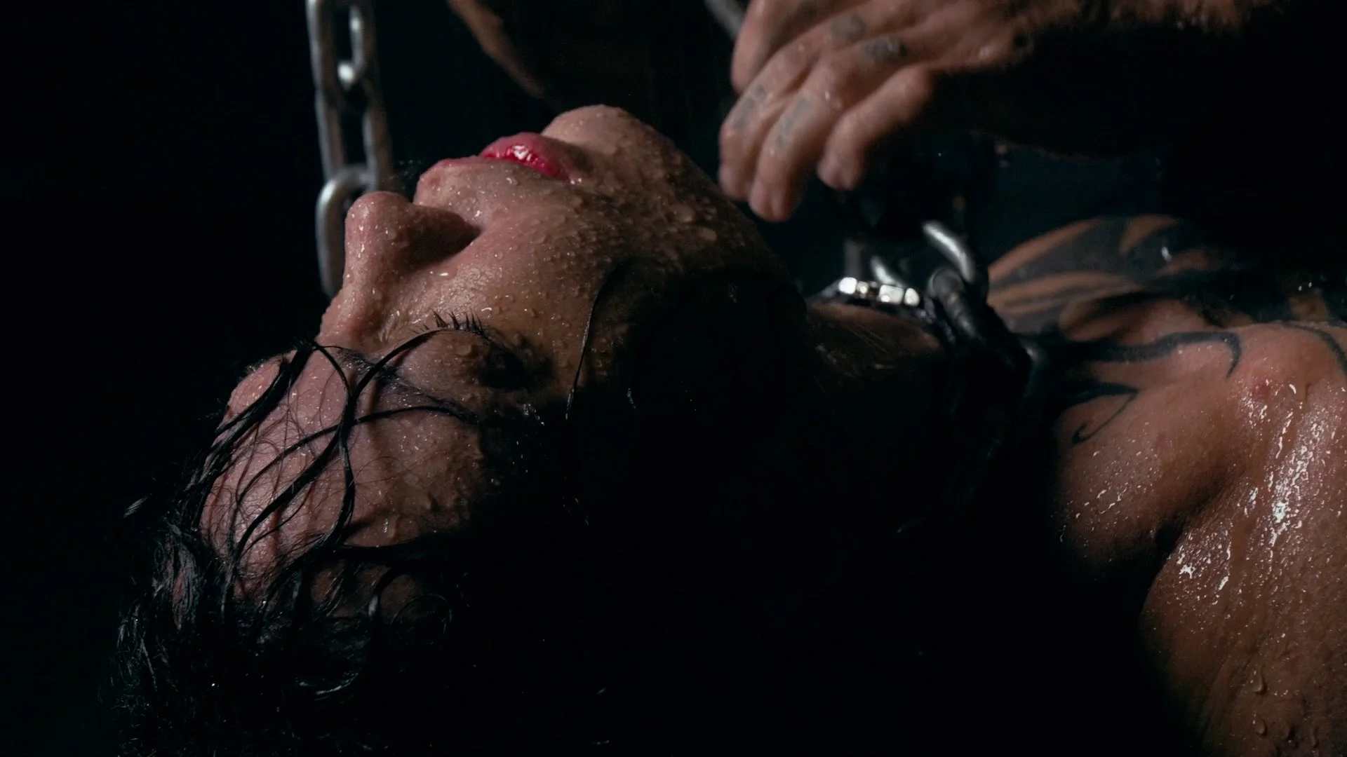 A close-up of a person with wet skin lying down, with closed eyes and lips slightly parted, water droplets on their face and body, and dark hair slicked back. Their hand is resting near their face.