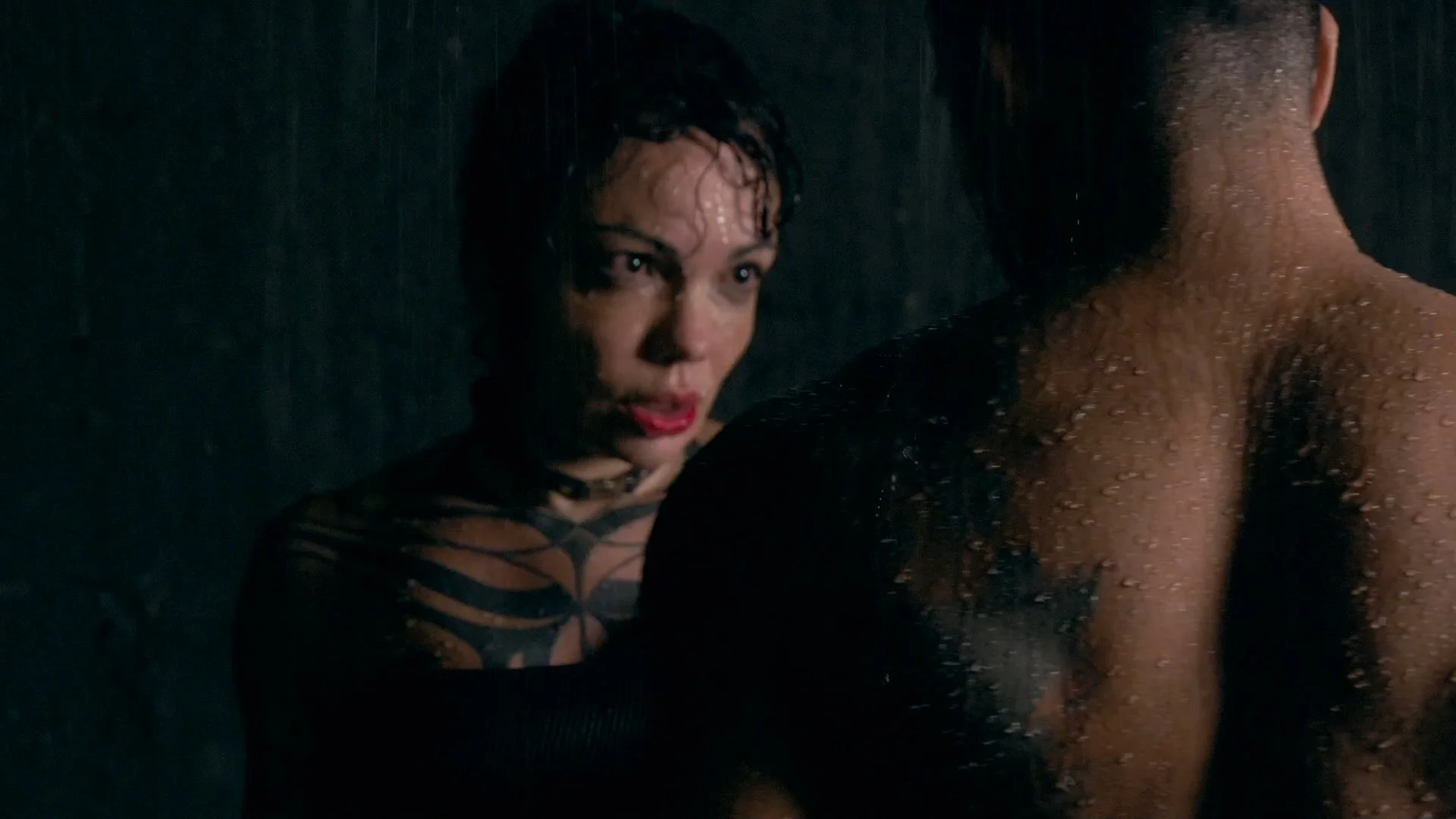 Woman with short curly hair and tattoos on her shoulder, looking at a shirtless man with wet skin, in a dark setting.