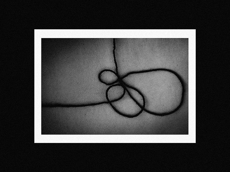 A piece of black string tied in a loose knot on a light gray surface, framed with a white border, against a black background.