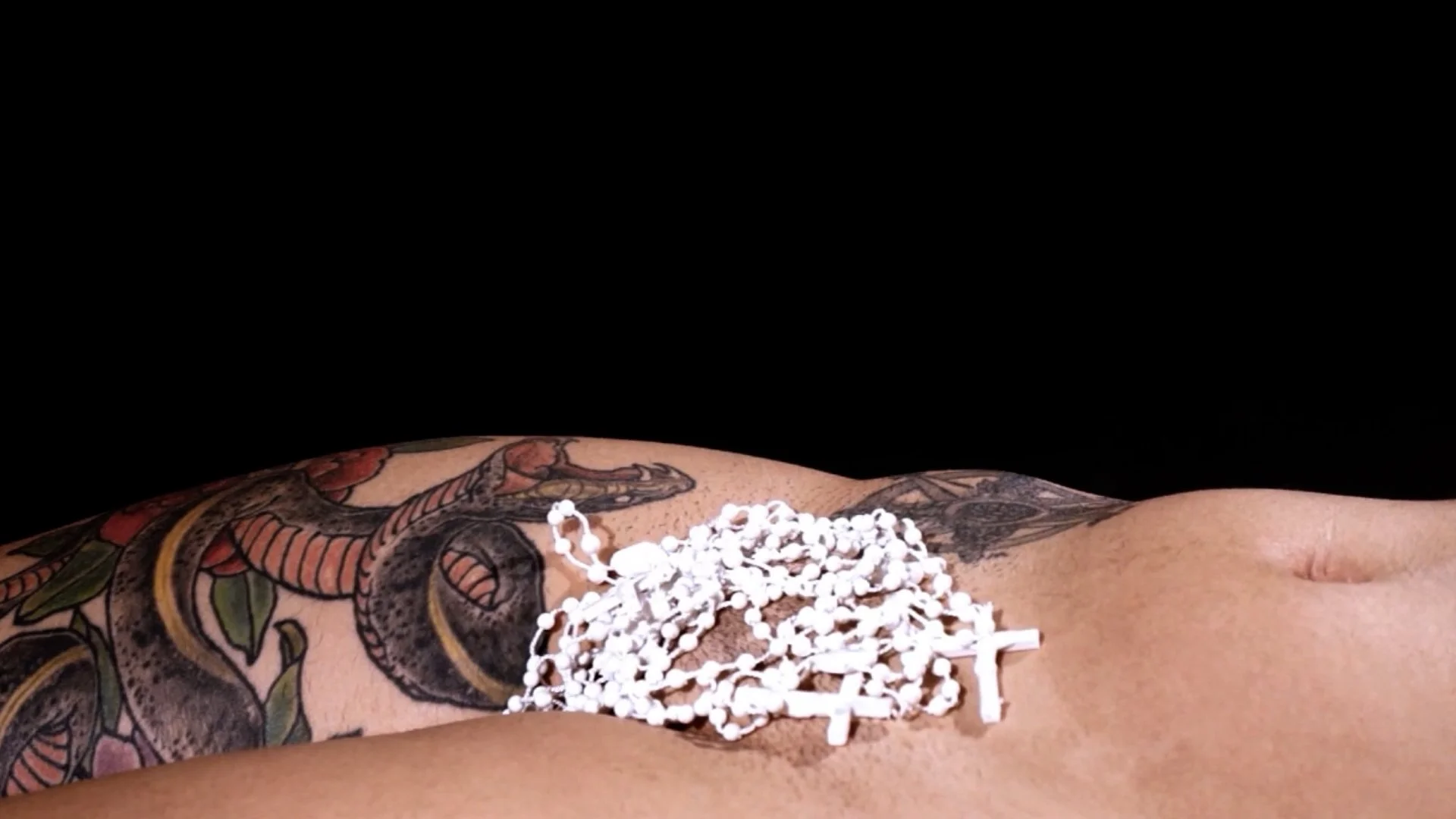 A person with tattoos on their side lying down, holding a white rosary with crosses on it, against a black background.