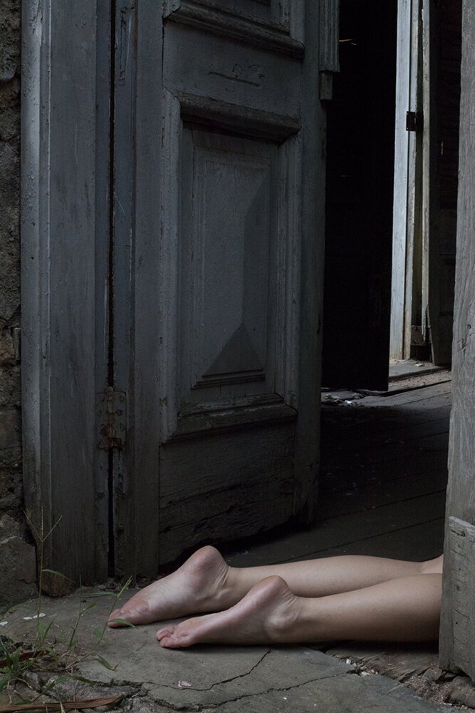 Close-up of human legs and feet laying on the ground outside an old, weathered wooden door with peeling paint, partially obscured by the doorframe.