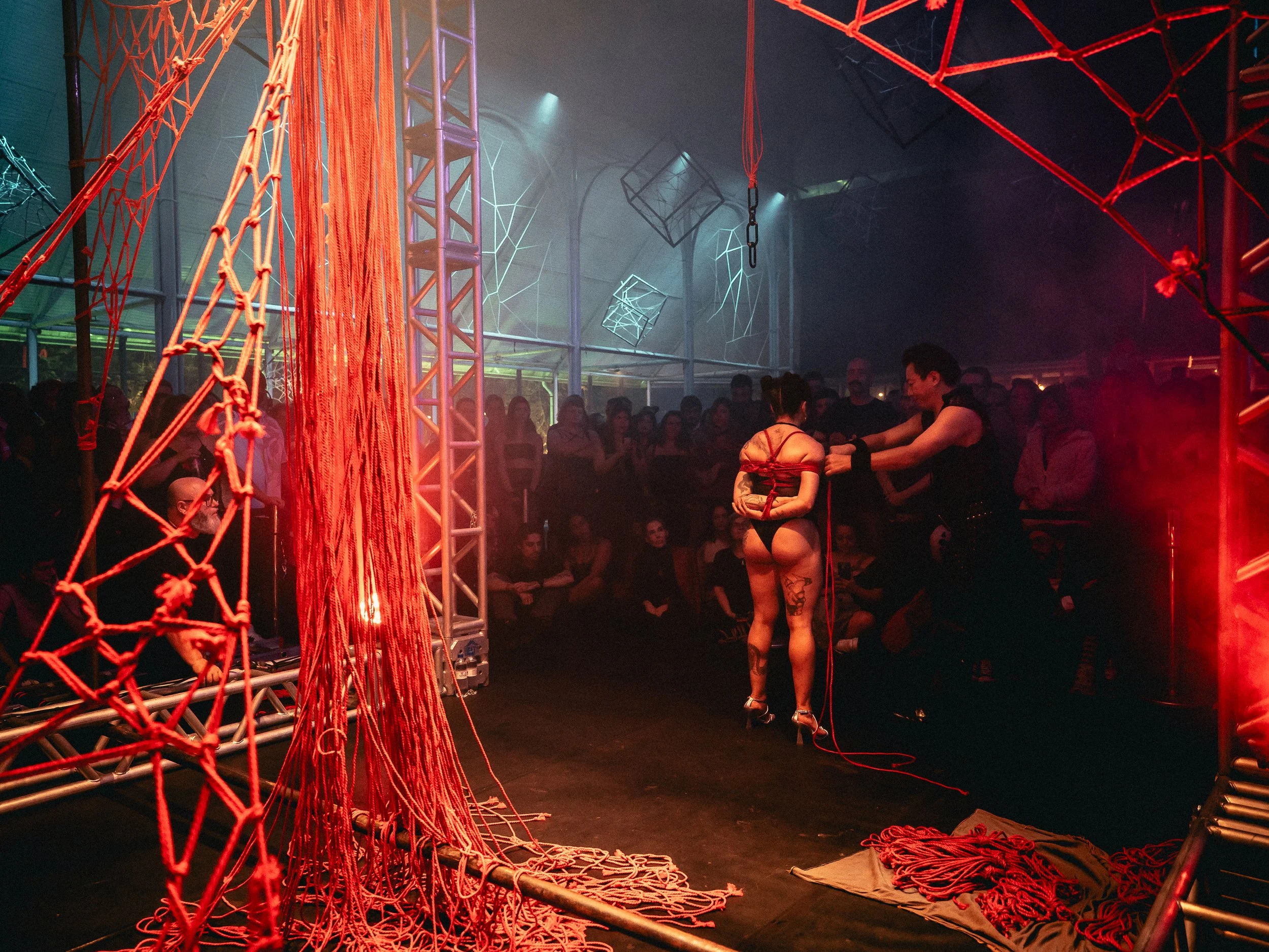 A staged performance in a dark, industrial-style venue with red and blue lighting. A woman in lingerie with tattoos is tied with ropes by a performer on a small stage, while an audience watches.