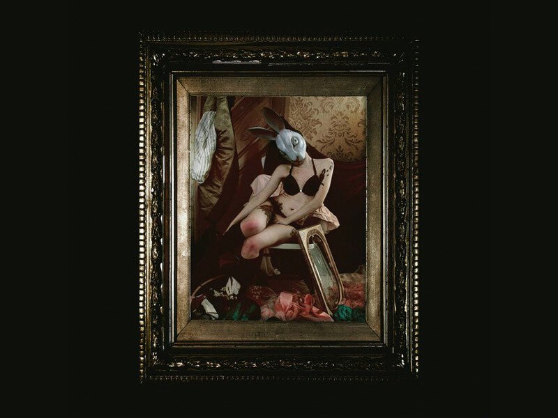 A person with a rabbit mask taking a mirror selfie in a dimly lit room, with a vintage frame mirror and various clothing items around.