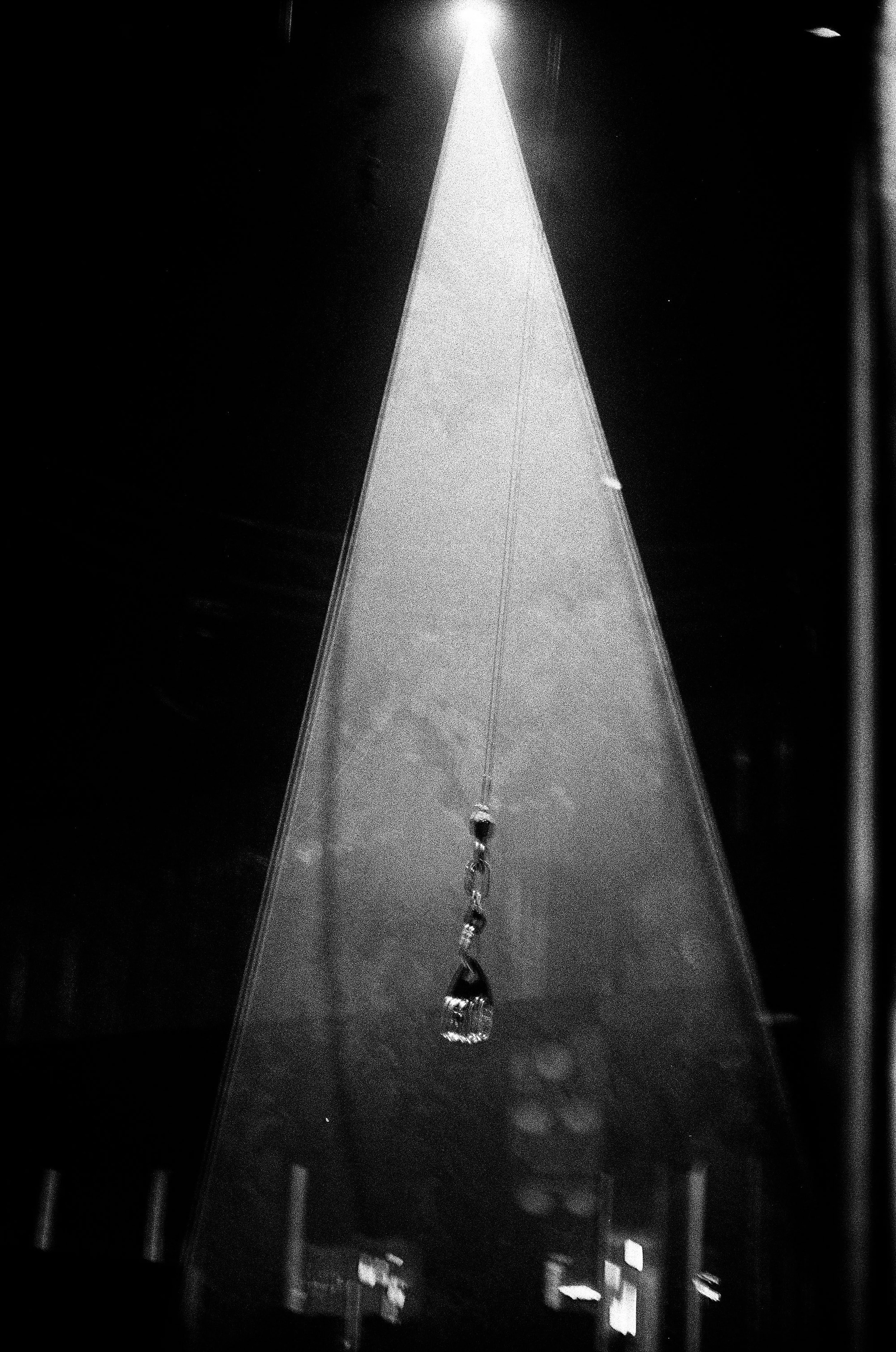 Close-up of a teardrop-shaped pendant hanging from a thin chain, illuminated by a bright overhead light in a dark background.