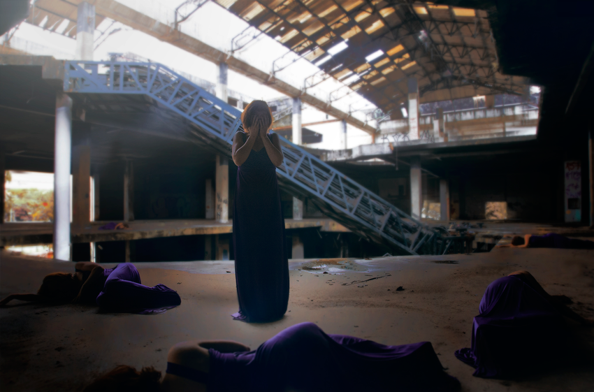A woman standing with her hands covering her face in an abandoned, partially destroyed building with debris and damaged roof structure, while others lie on the ground around her.
