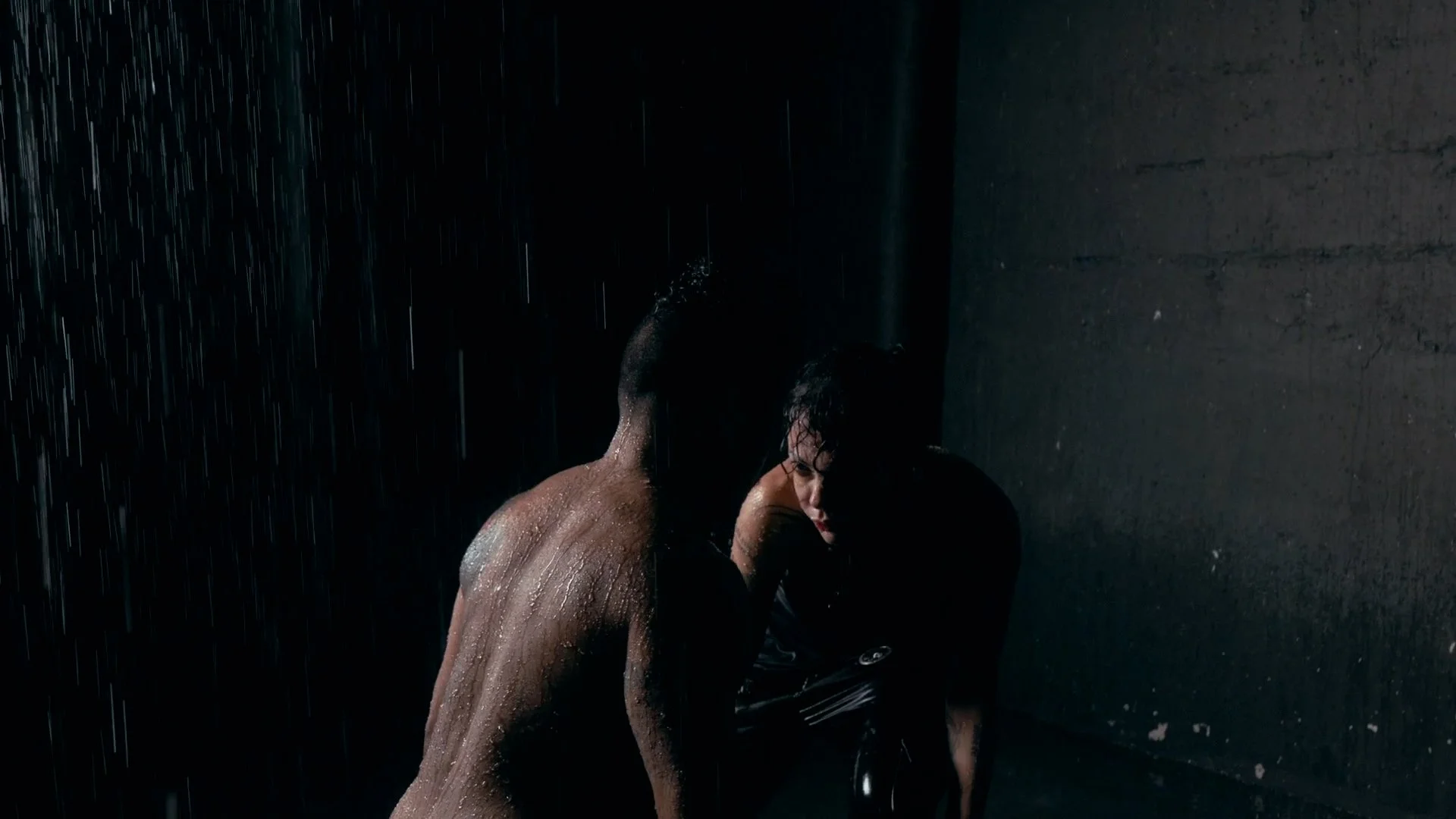 A man and woman in a dark room, wet from rain, with the man shirtless and the woman holding a black umbrella.