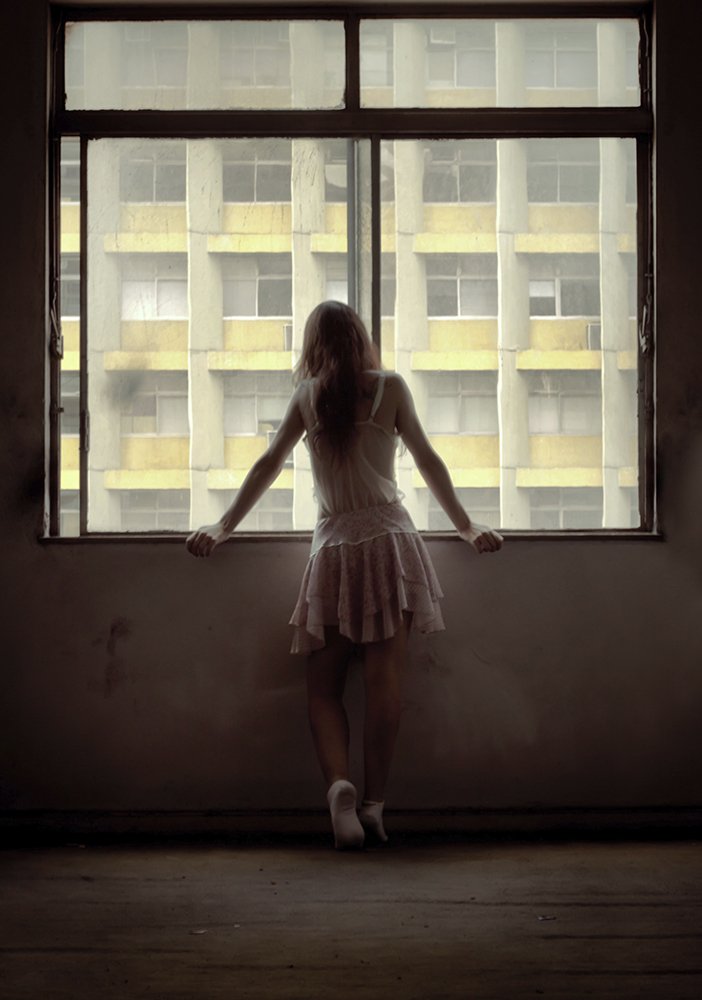 A woman in a white dress standing by a window, looking out at a city building with balconies.