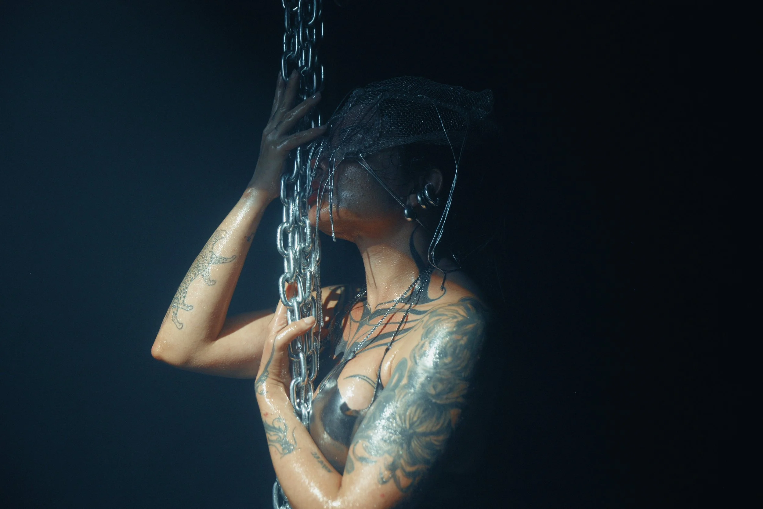 A woman with tattoos on her arms and chest, wearing a black head covering and earrings, holding onto a metal chain in a dark setting with water on her skin.