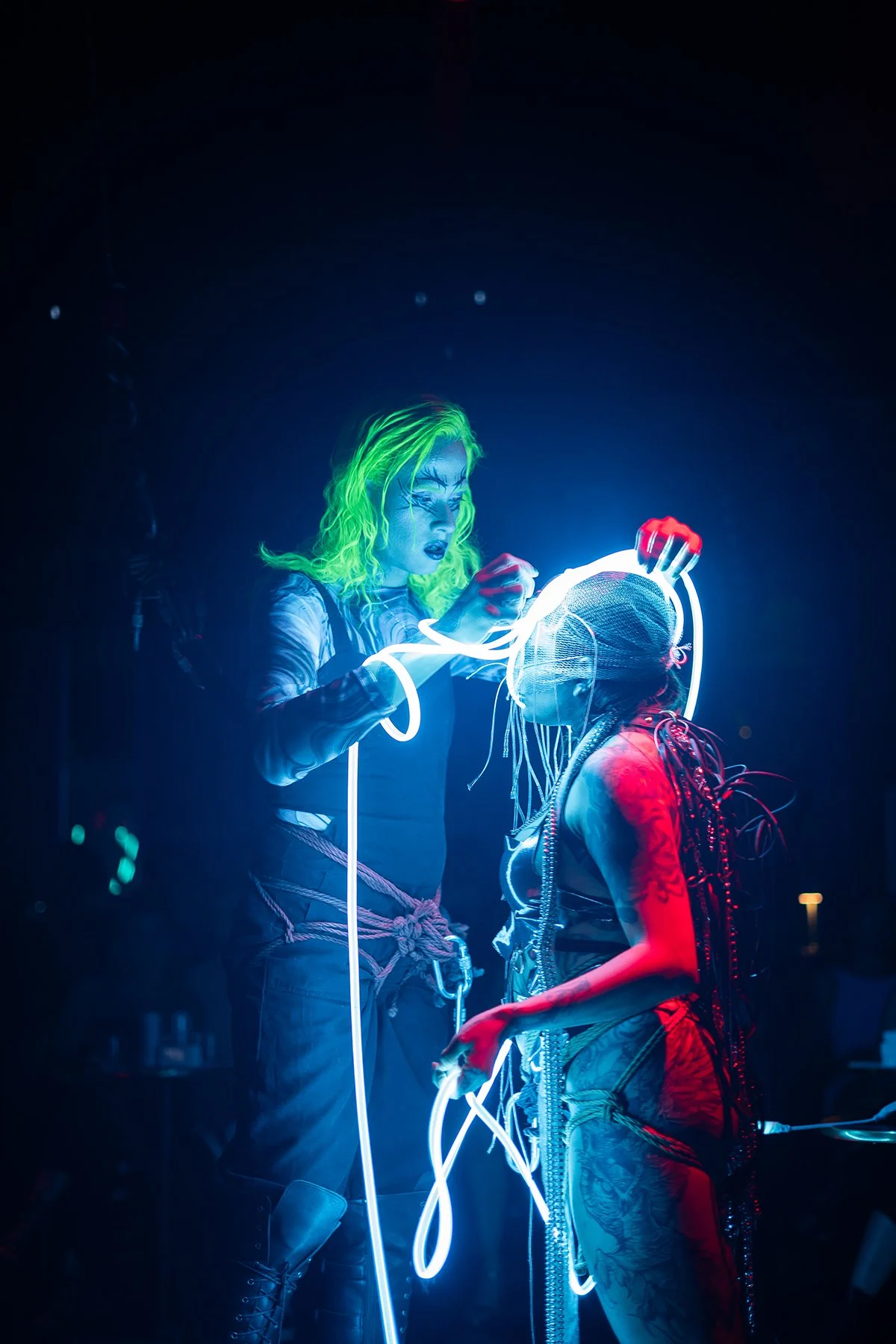 Two performers engaged in a futuristic neon-lit performance with glowing wires, one with neon green hair and makeup, the other with blonde braids and tattoos, in a dark setting.