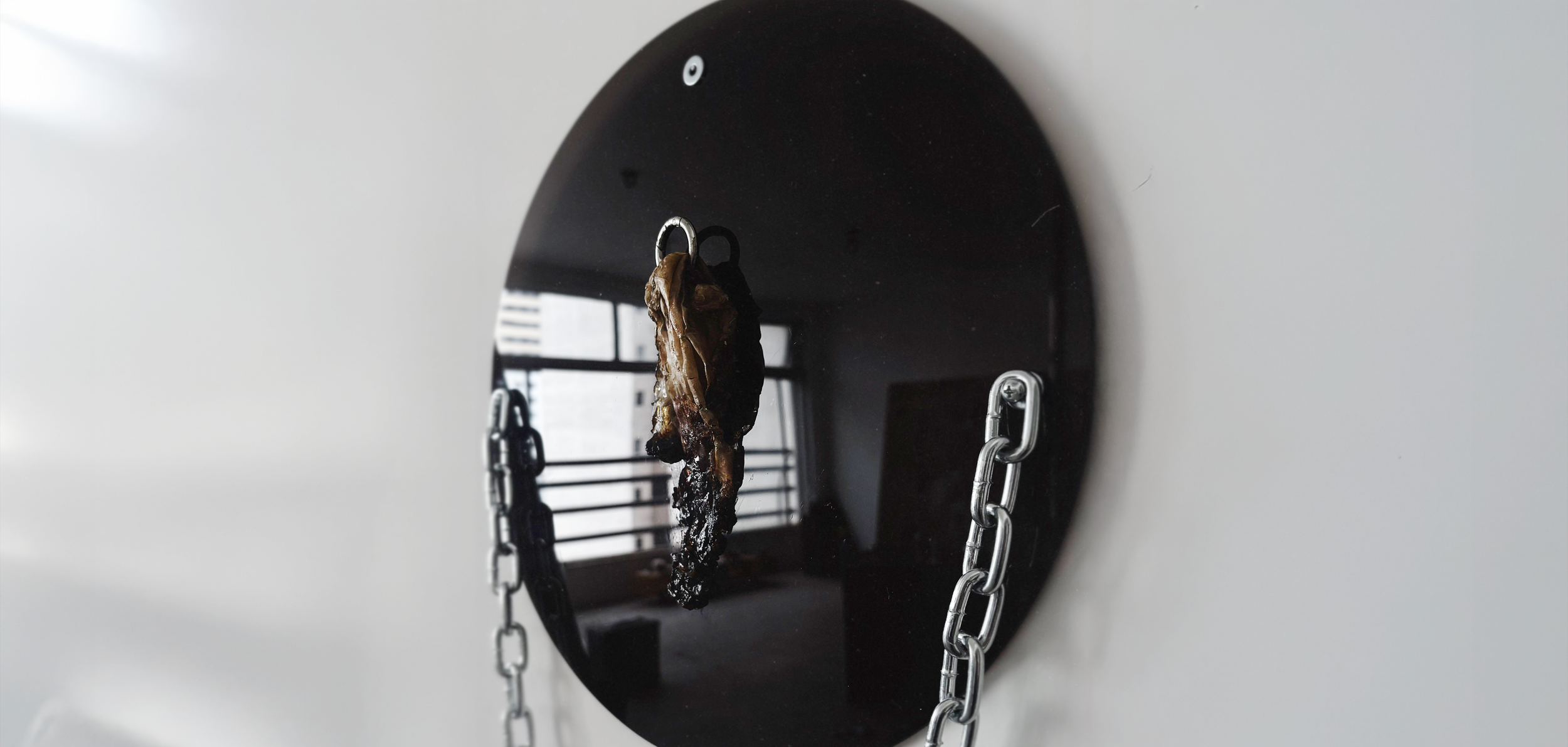 Charred piece of meat hanging from a mirror with chains, inside a room with windows in the background.