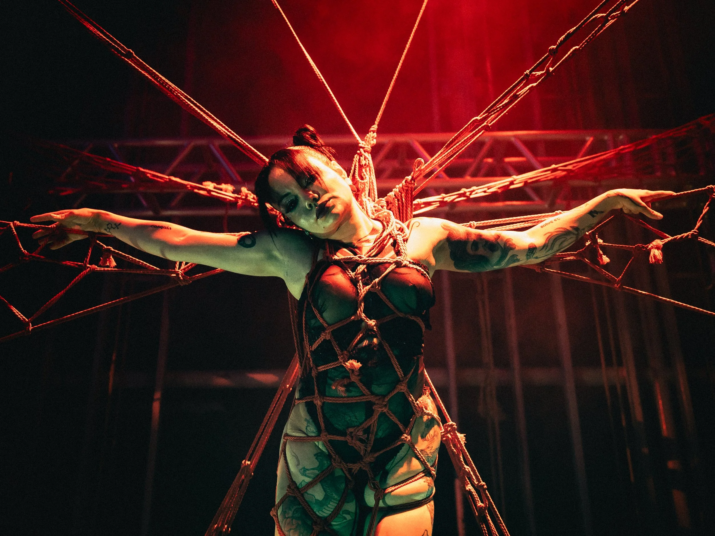 A woman with tattoos on her arms and dark hair in a bun stands with her arms outstretched in a web of ropes, illuminated by red lighting.