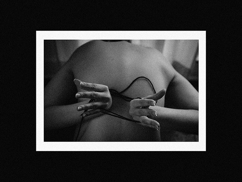 Black and white photo of a person lying down with hands extended, wearing a thin strap lingerie or clothing, with a white border frame.