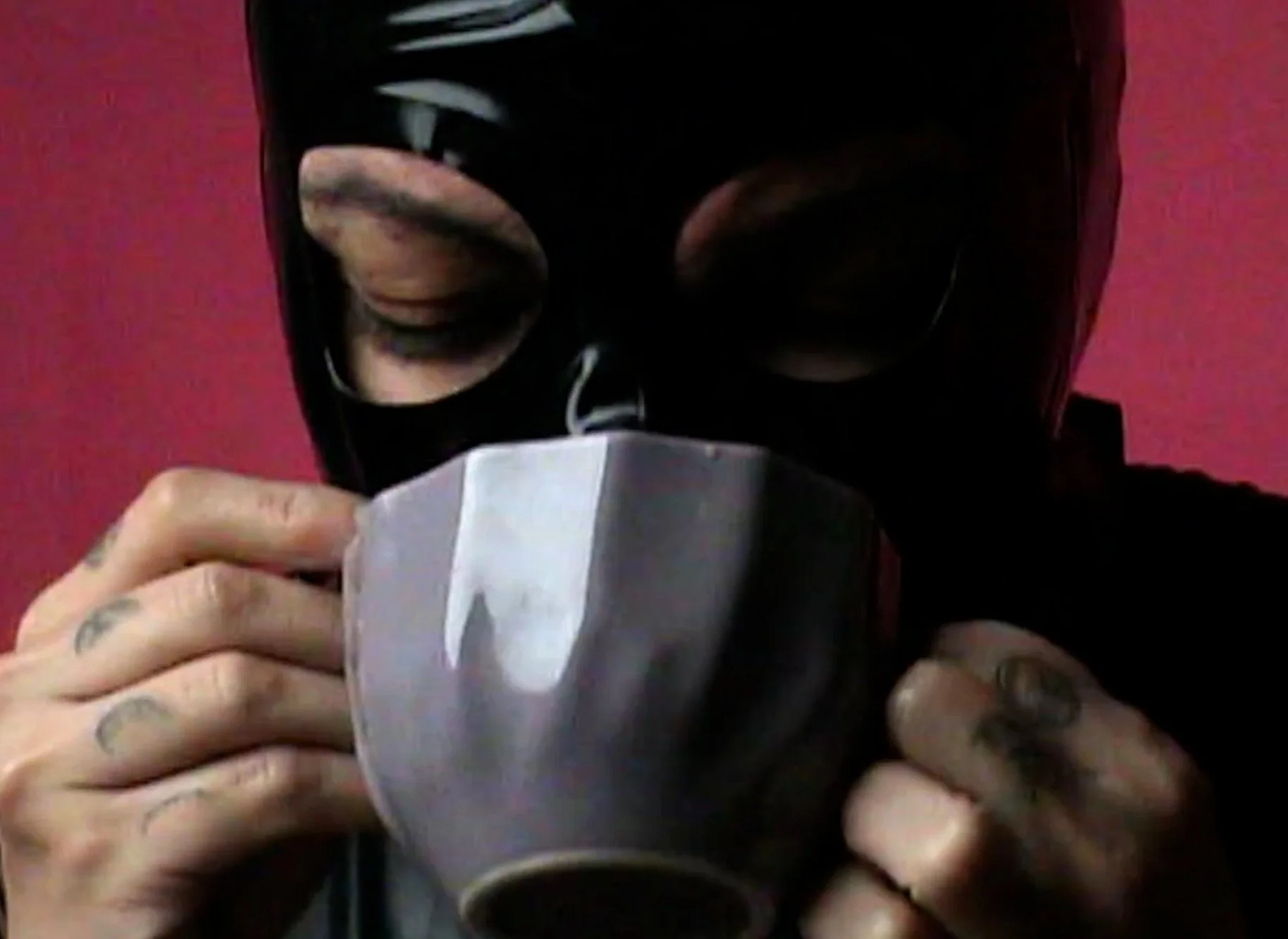 Person wearing a black mask and black clothing, holding and drinking from a gray mug, against a red background.