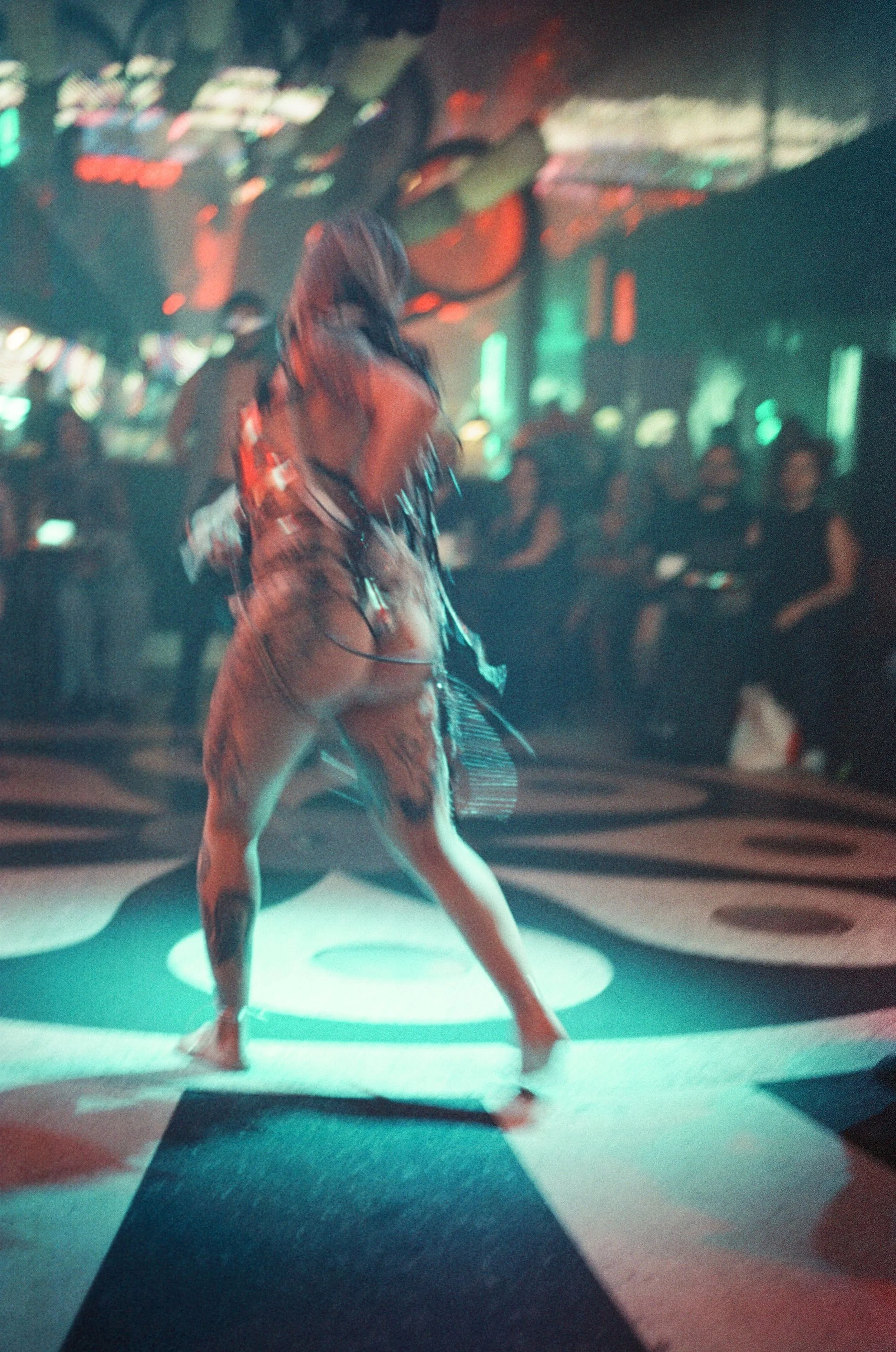 A woman with long hair, tattoos, and wearing a crop top and shorts dancing barefoot on a circular stage, illuminated by teal and red lights, with an audience in a dimly lit nightclub in the background.