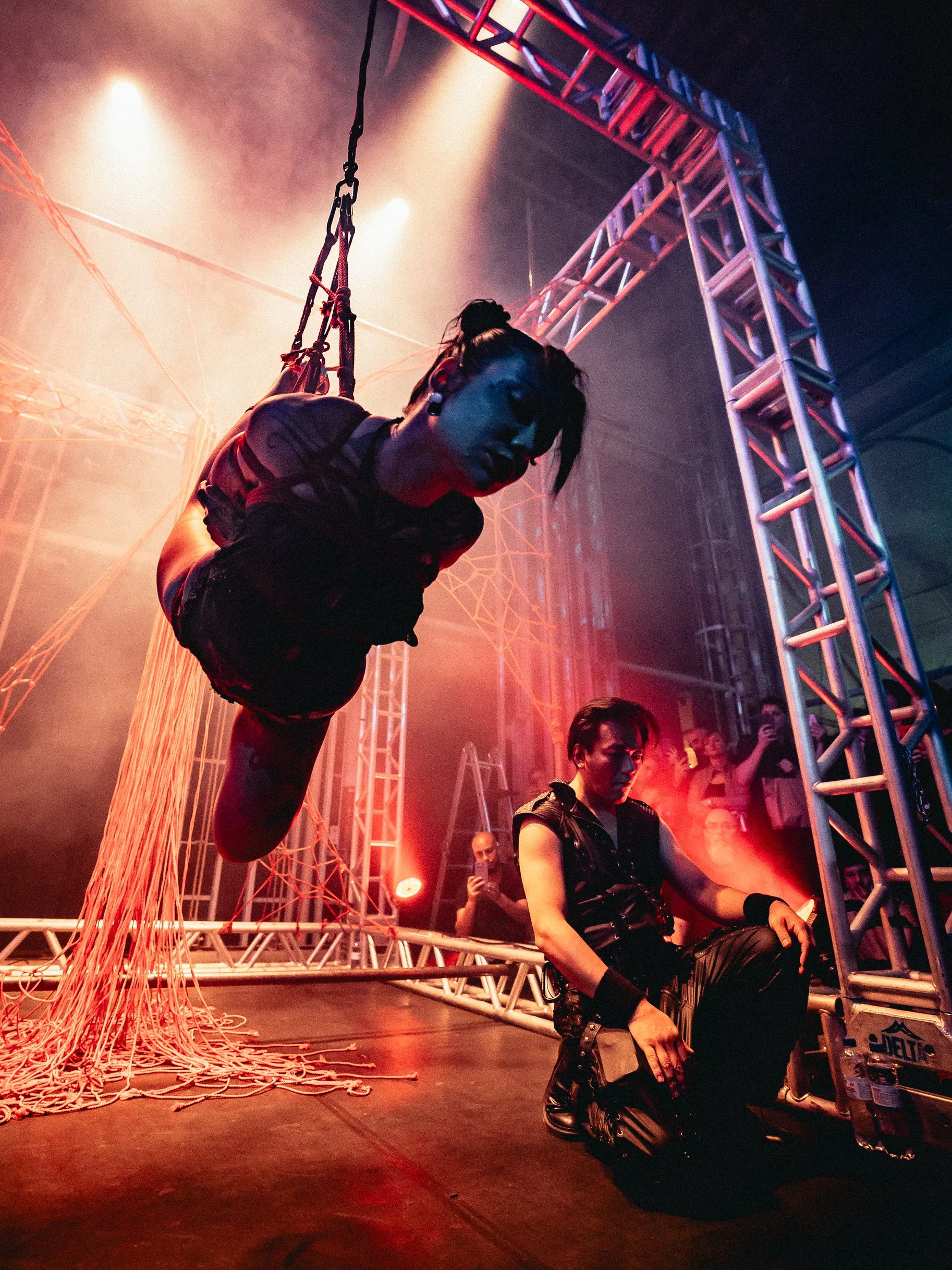 A performer suspended in mid-air on a trapeze, with a person kneeling on the stage below, at a circus or performance event with colorful stage lighting and audience members in the background