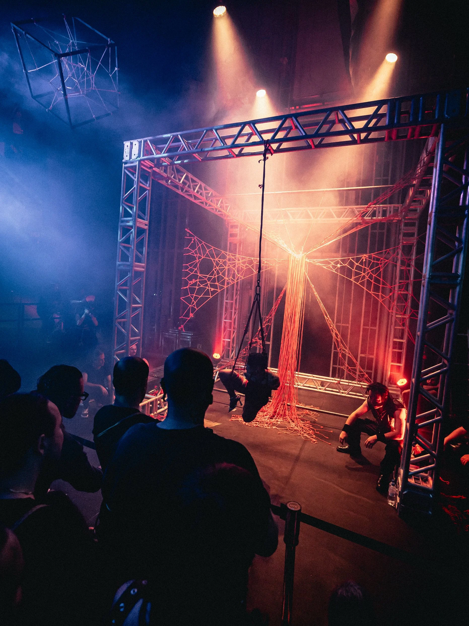 A performer swinging on a trapeze inside a metal cage structure decorated with orange web-like yarn, with stage lighting and an audience watching.