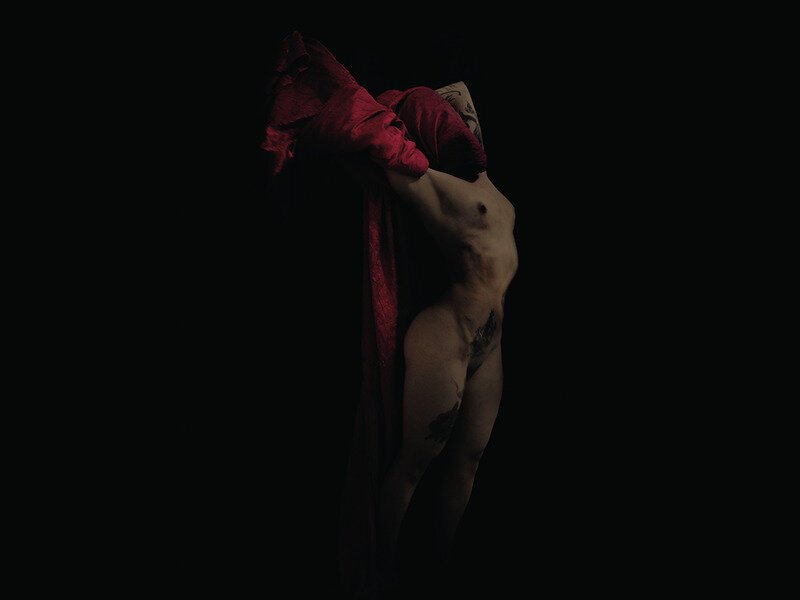 A person is partially covered with a red fabric, with their body exposed and visible tattoos, standing in a dark environment.