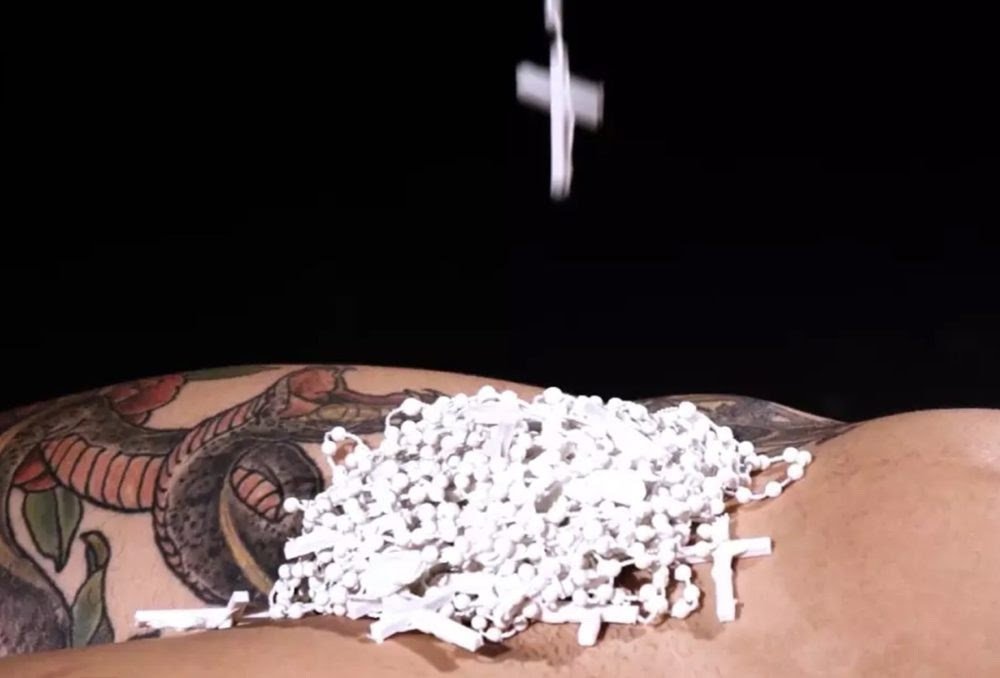 Person pouring small white pills or tablets onto their tattooed arm, with a cross tattoo visible.
