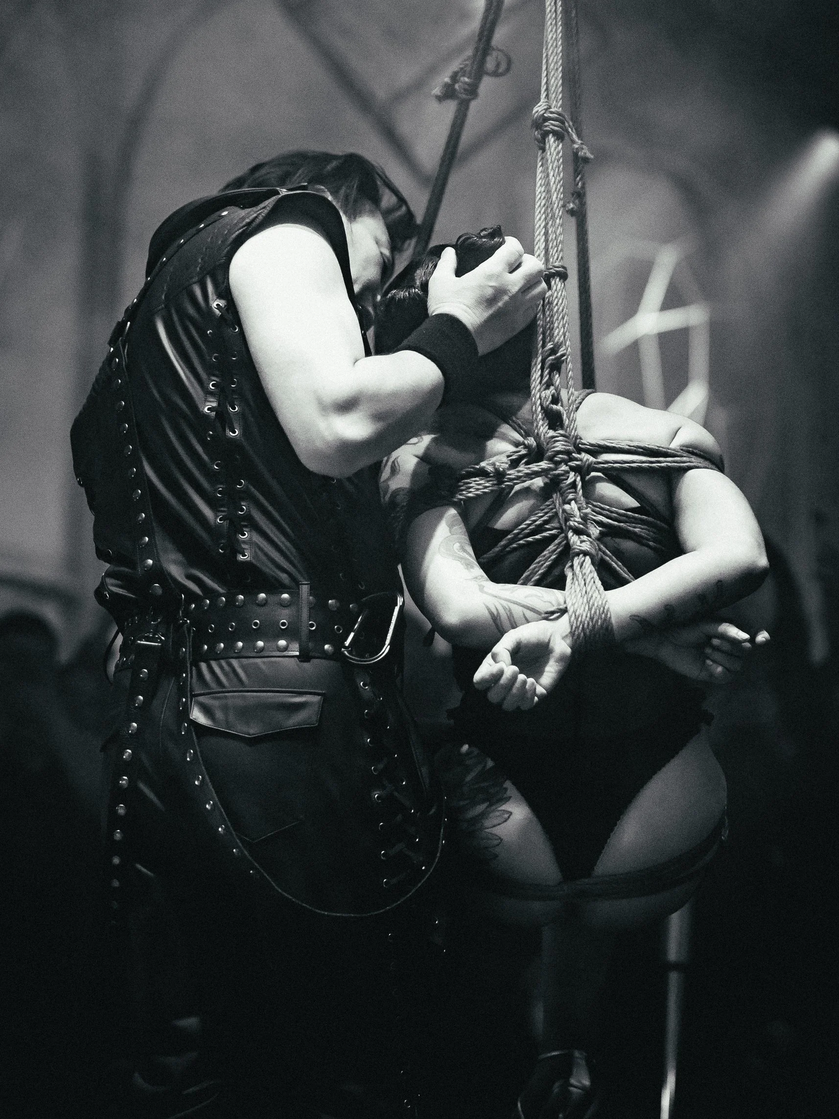 A person dressed in black leather clothing kisses a tied-up and gagged woman during a BDSM scene in a dark room.