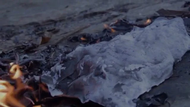 Close-up of a charred paper or fabric on a fire with ashes and burnt debris around.