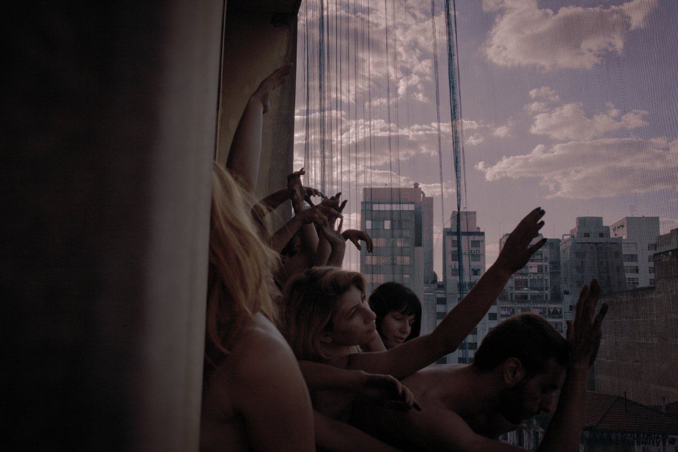 A group of people with raised hands and expressive body language sitting by a window with city buildings in the background, during what appears to be a sunset or sunset-like lighting.