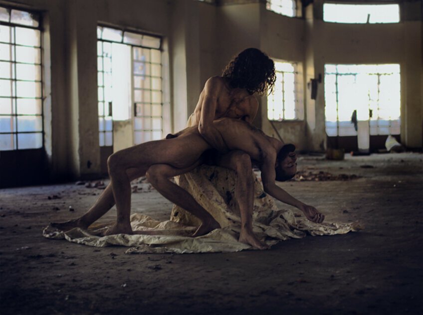 Two people engaged in an intimate pose on a sheet inside a dilapidated building with large windows and debris on the floor.