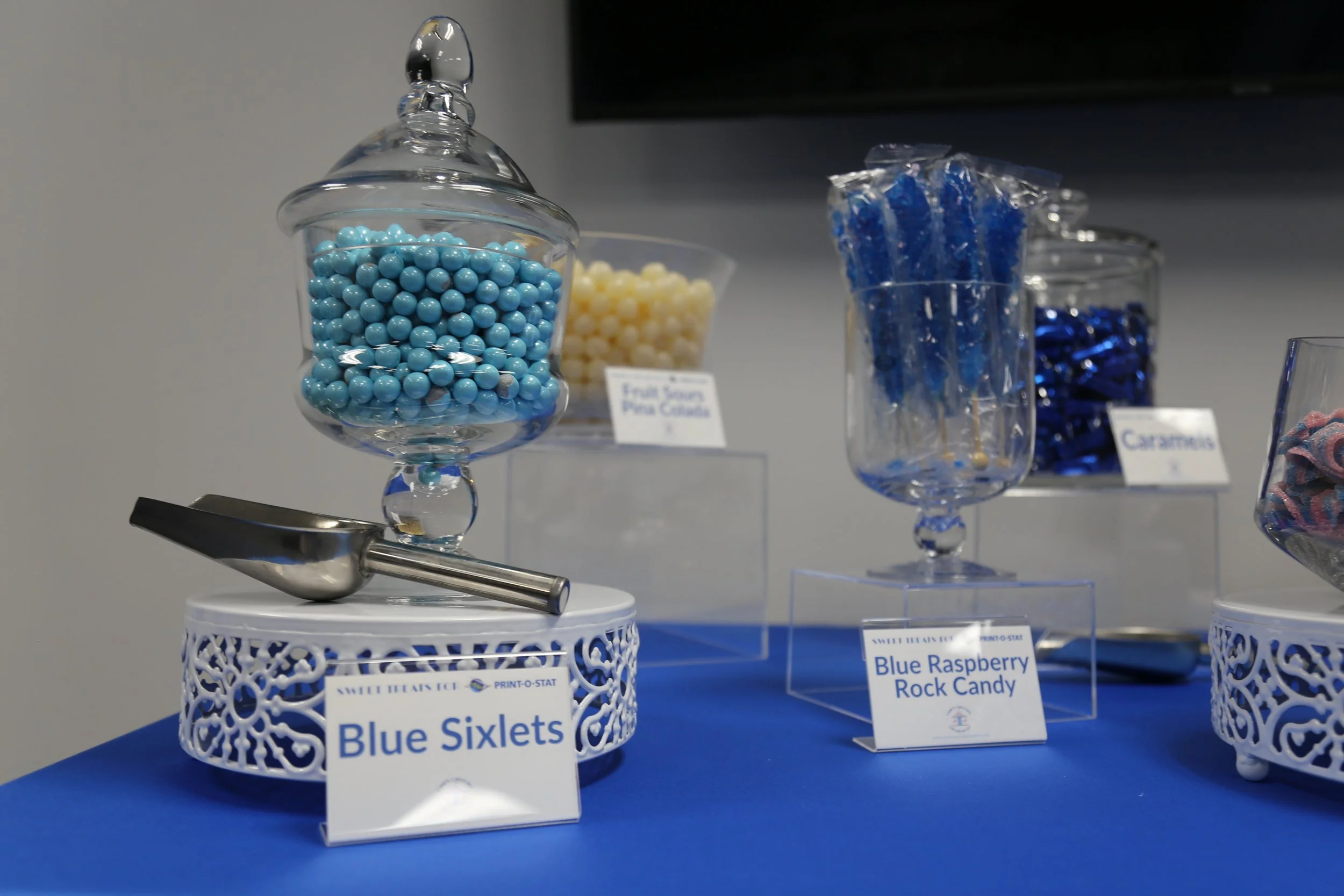 Display of blue and white candy, labeled as Blue Sixlets and Blue Raspberry Rock Candy, in decorative glass jars on a blue table.