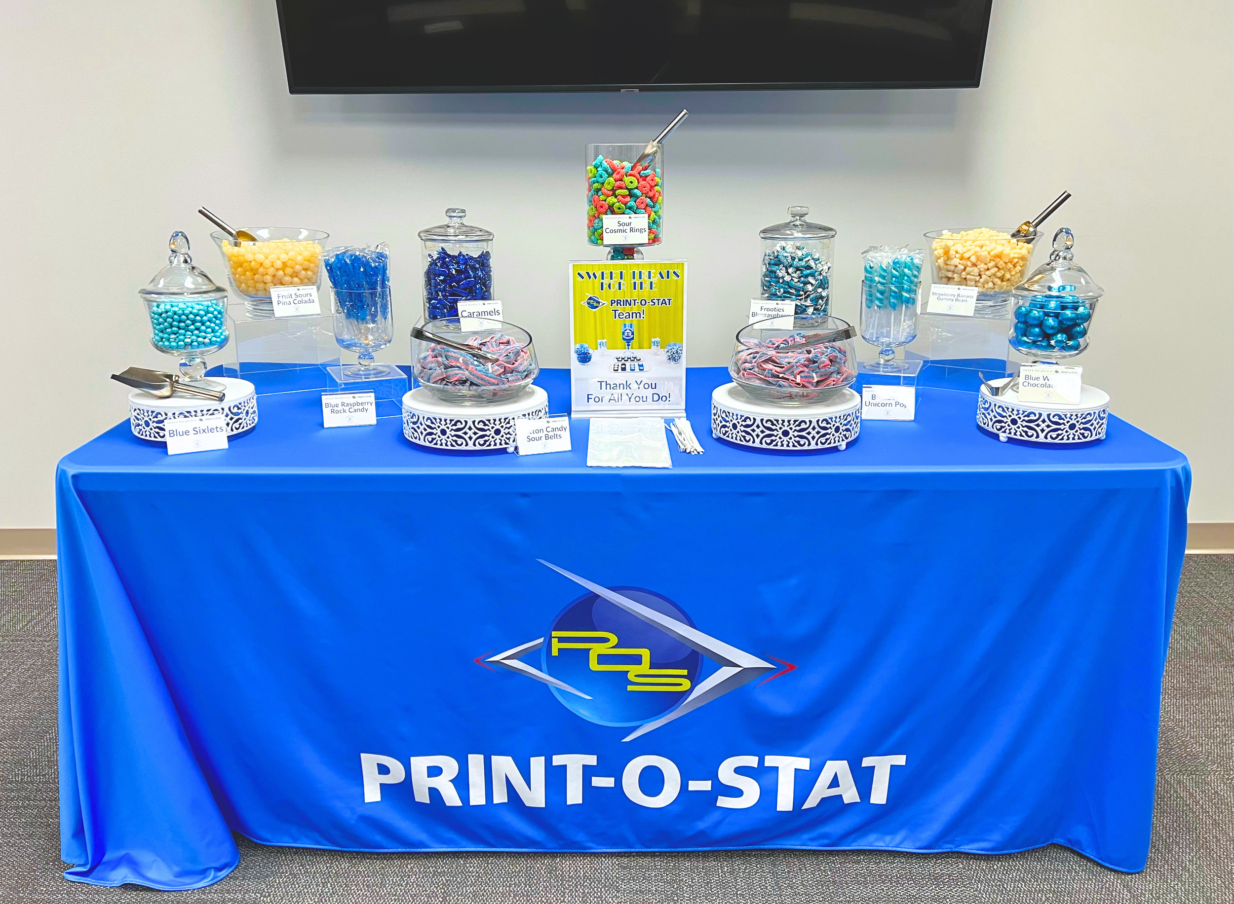 A table covered with a blue cloth displays an assortment of colorful candies in glass jars. Each jar is labeled with the candy name, such as Blue Sixlets, Fruit Sours Pina Colada, Caramels, Sour Cosmic Rings, Footies Blue Raspberry, Blue Unicorn Pop, and Blue W Chocolate. A sign in the center reads "Sweet Treats for the Print-O-Star Team!" and "Thank You For All You Do!" There is a larger jar of Sour Cosmic Rings in the middle with a small scoop, and other jars with scoops as well.