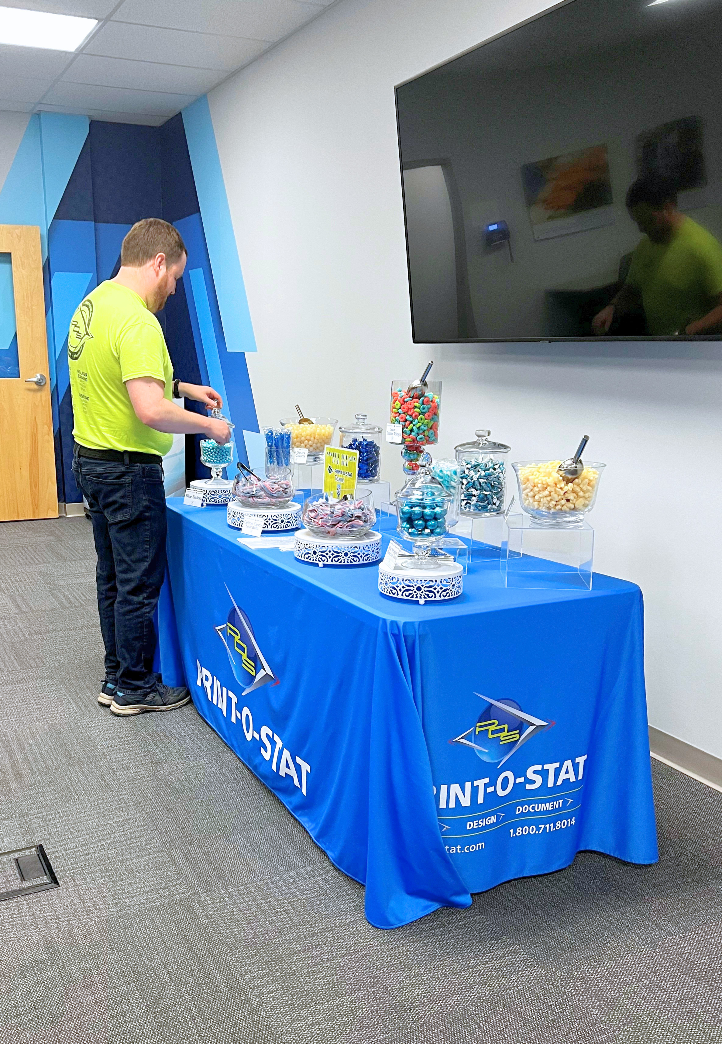 A man in a yellow T-shirt with a logo is standing at a table covered with a blue cloth, arranging colorful candies in jars at an event. The table has a logo and text that reads 'PRINT-O-STAT' and contact information. There is a large flat-screen TV mounted on the wall behind the table, and a framed picture and a thermostat can be seen on the wall. The room has a carpeted floor and a ceiling with rectangular lights.