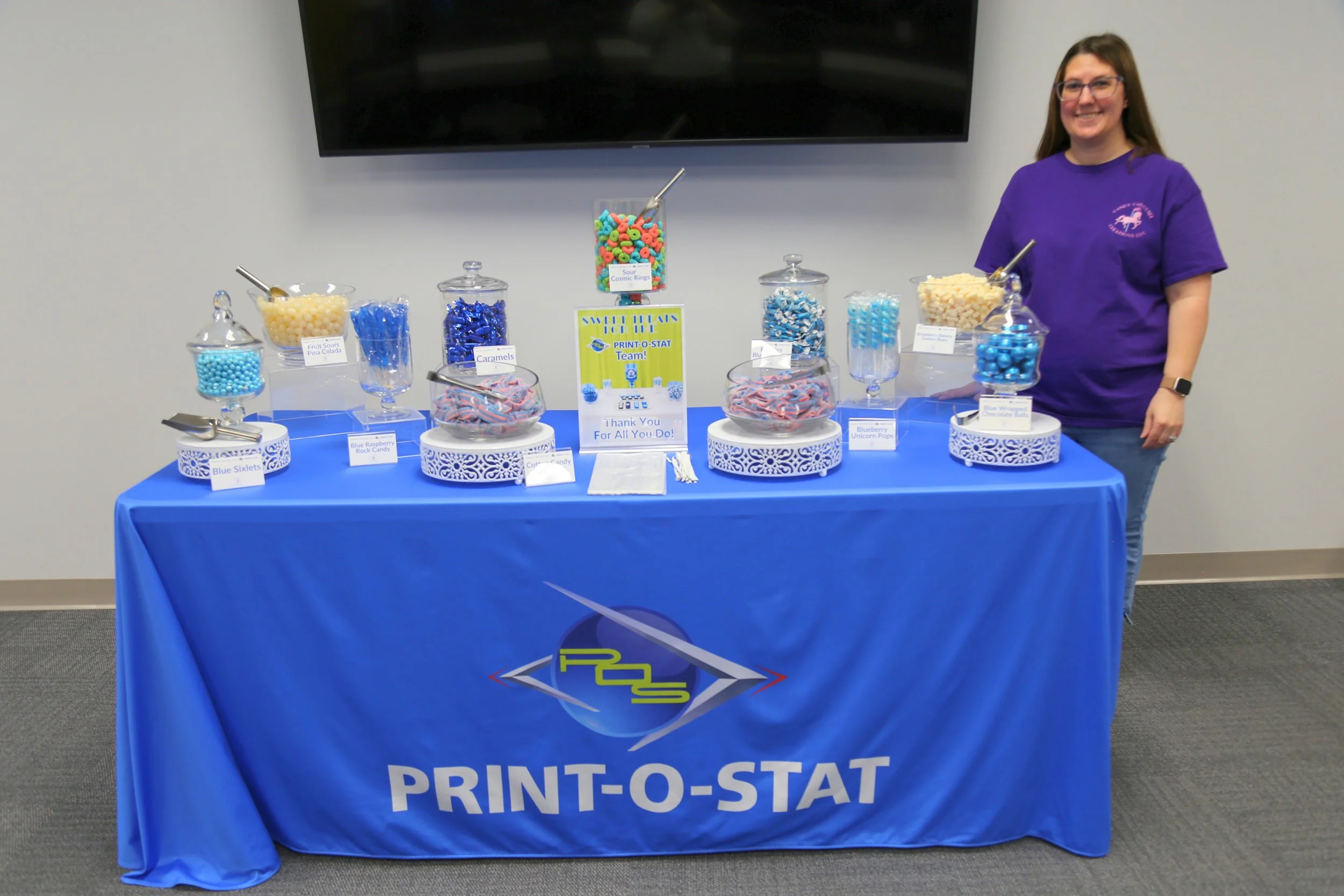 A woman in a purple shirt standing behind a table covered with a blue tablecloth at a school event. The table displays various colored candies in glass jars with labels, including blue sixlets, fruit sours, sour cosmic rings, candies, caramel candies, blue raspberry rock candy, and icy blue candies. There is a sign on the table that reads "Thank you for all you do!" and a large TV screen is mounted on the wall in the background.