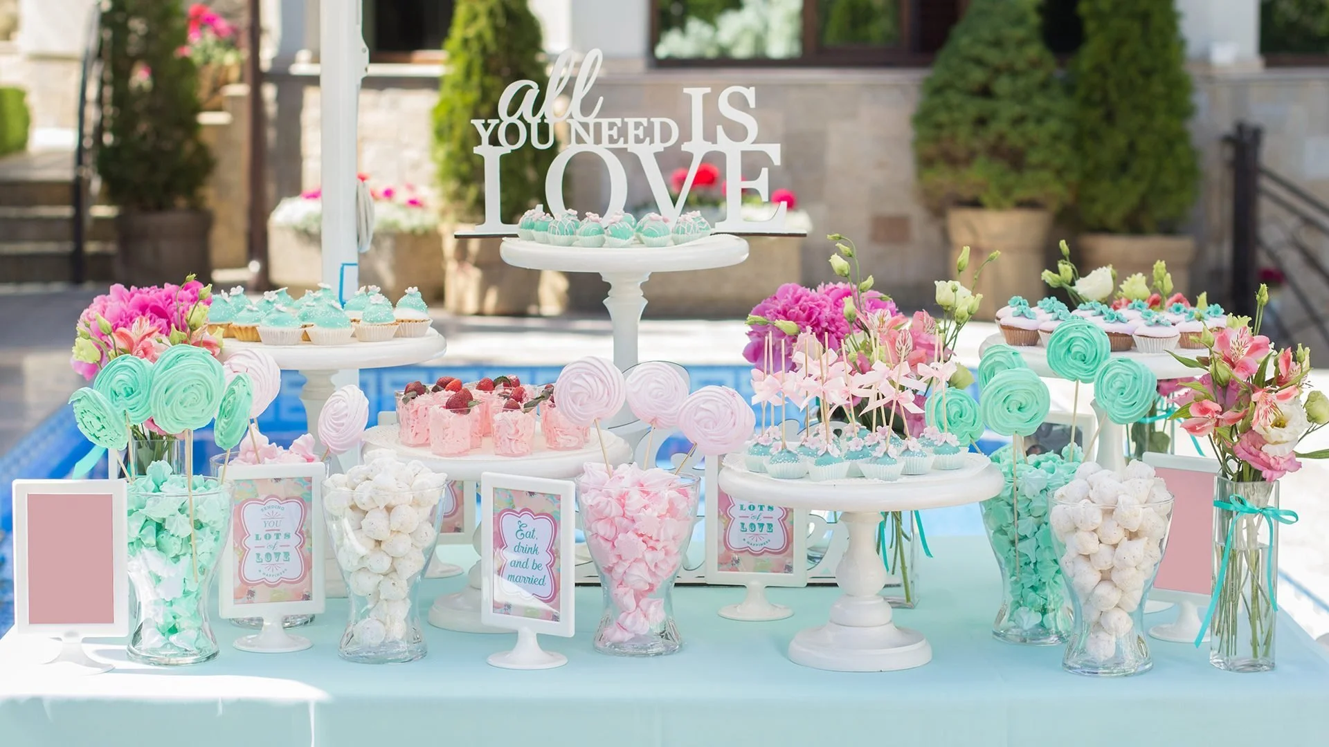 A pastel-colored dessert table with cupcakes, pink and blue cake pops, and confections, decorated with pink flowers, green lollipops, and framed signs, set outdoors near a pool with potted plants in the background.