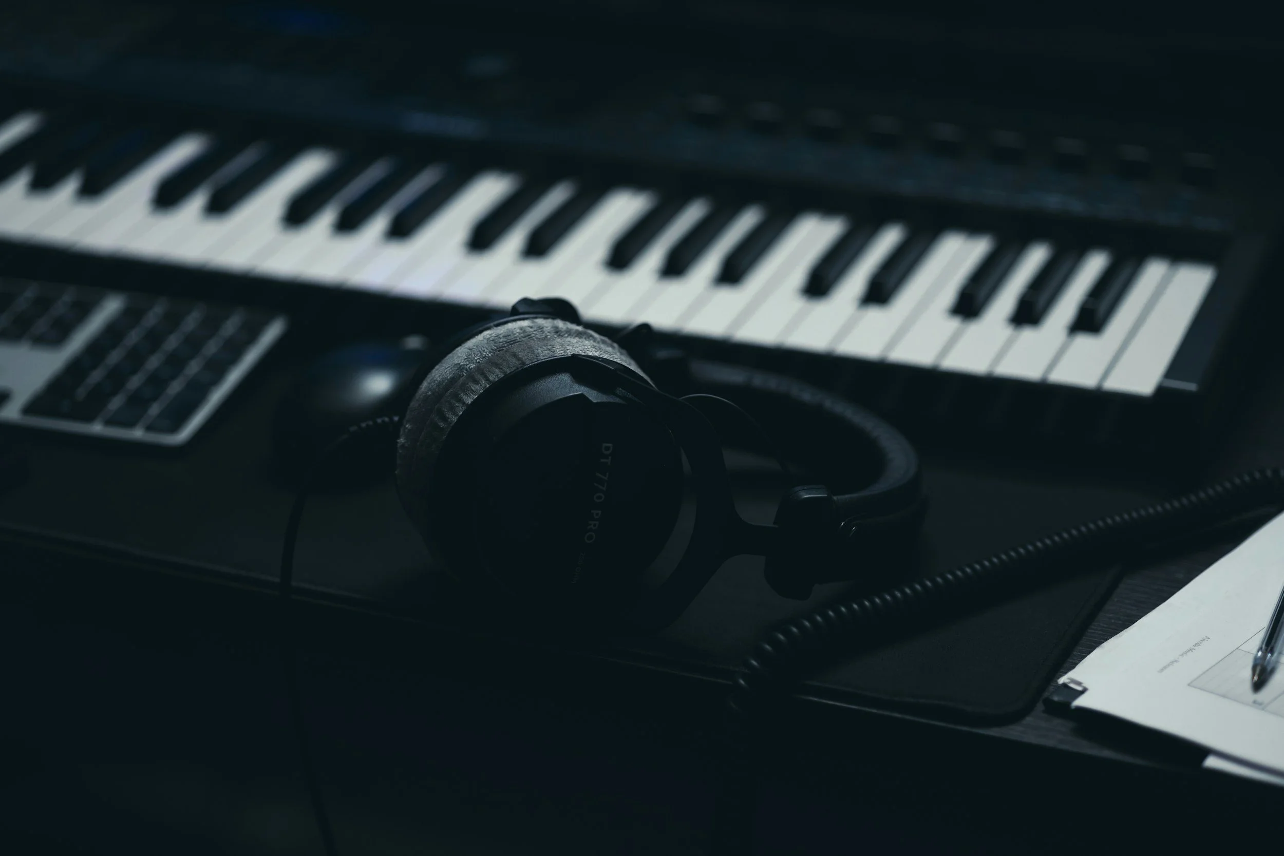 A pair of black headphones resting on a black laptop next to a keyboard and a notepad with a pen.