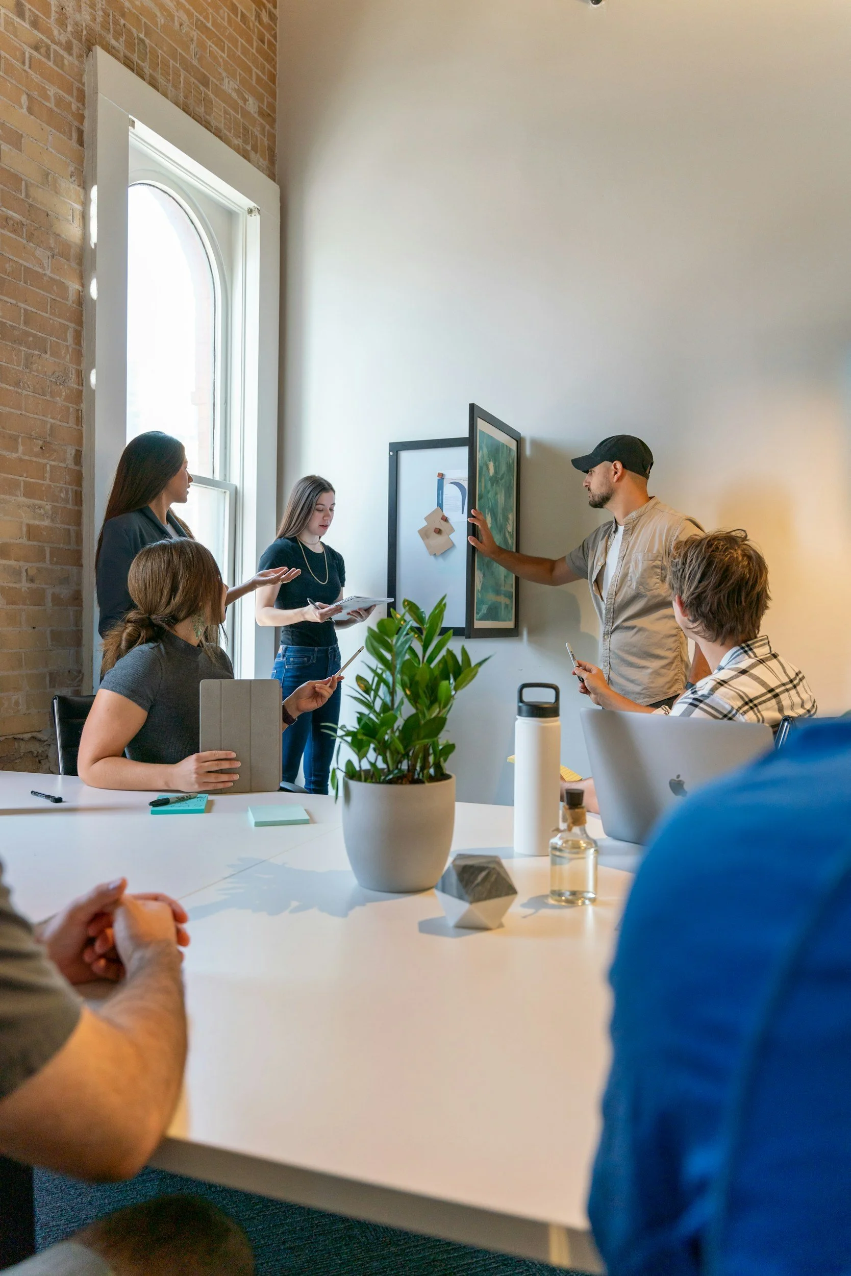 Group of five people in a meeting room with a white table, a potted plant, and a whiteboard, engaged in discussion. One woman stands near the whiteboard, while others sit around the table with laptops, tablets, and notebooks.
