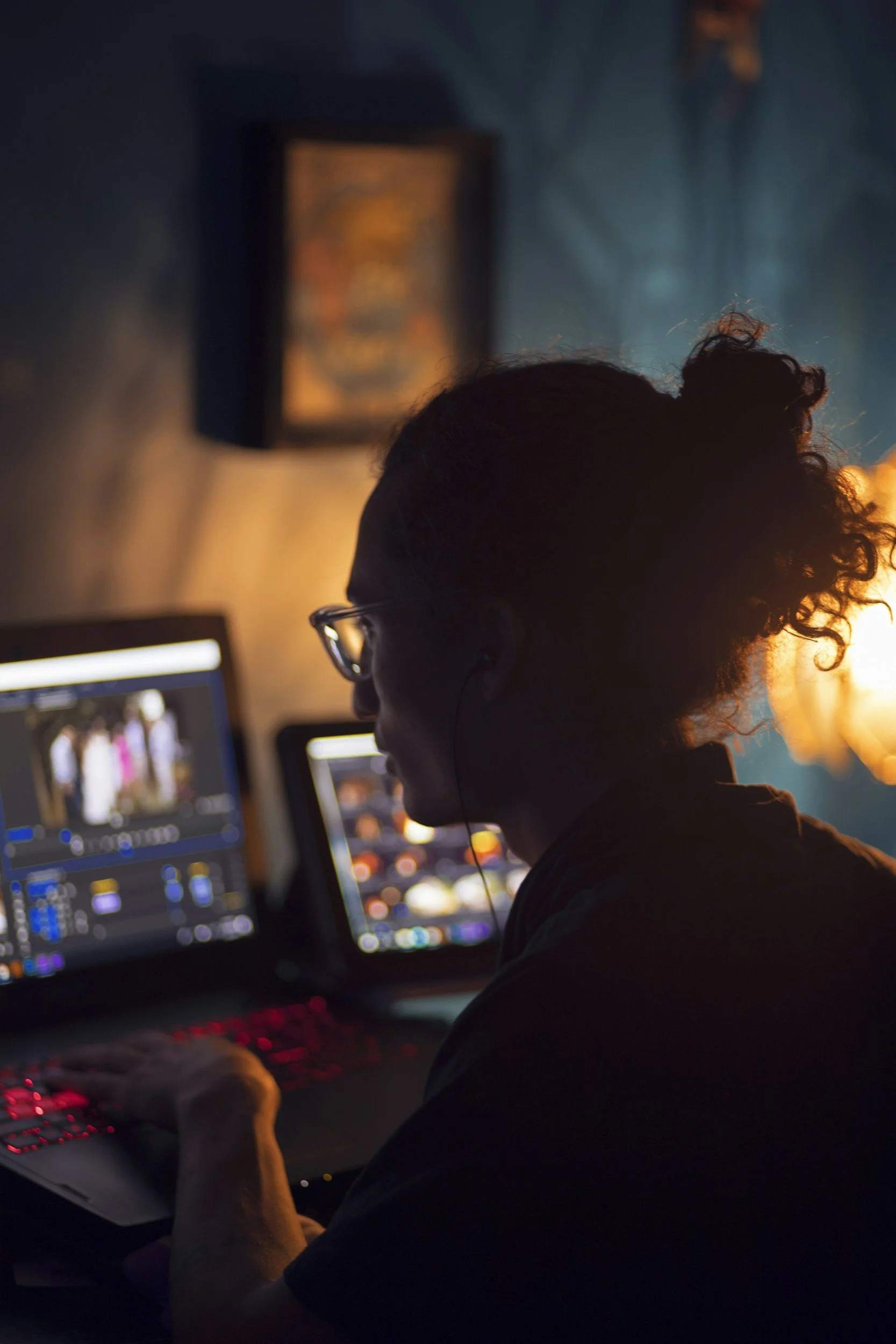 A person with curly hair and glasses working on video editing on a computer in a dimly lit room.