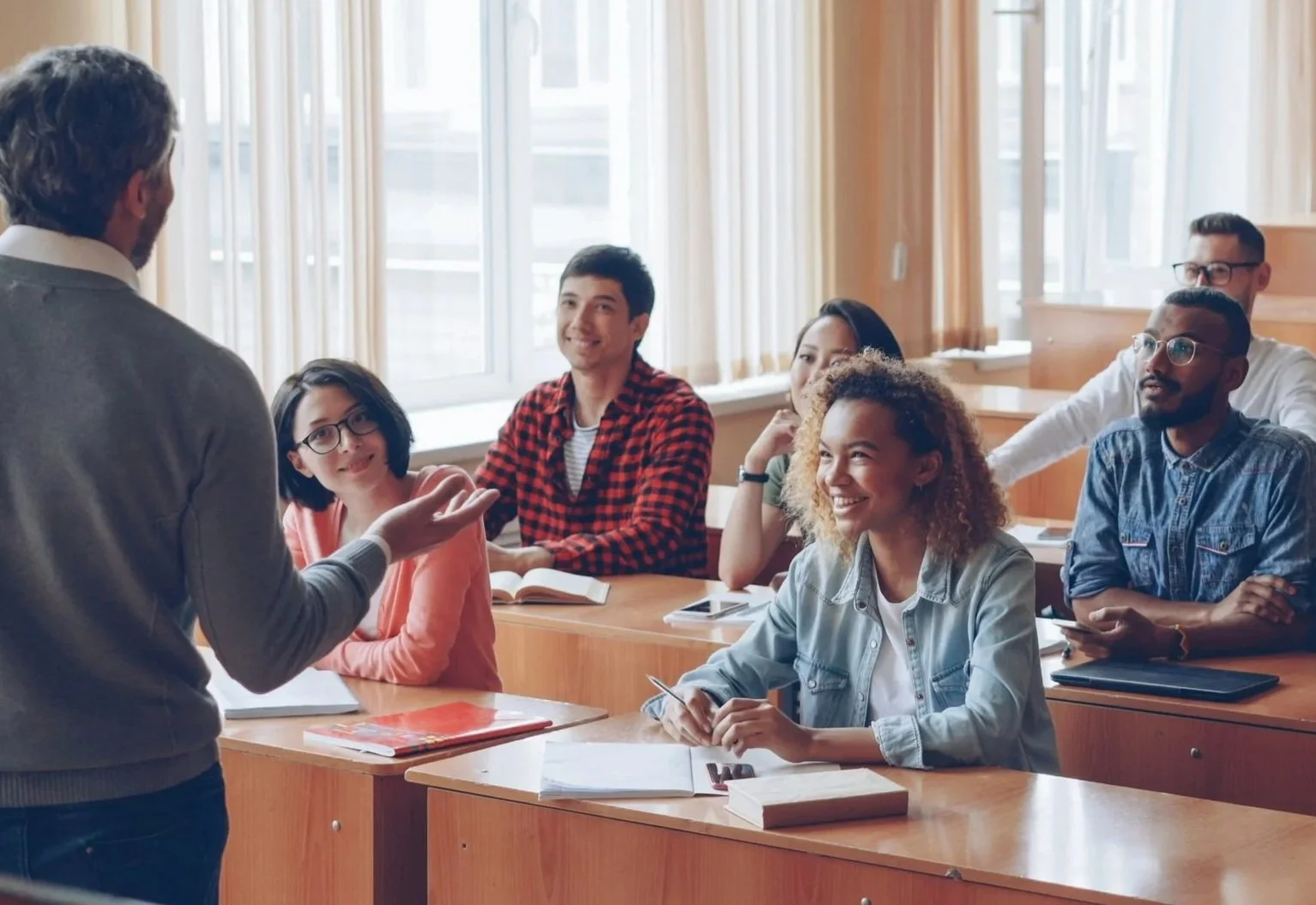 Professor speaking to students in a classroom with large windows and wooden desks.