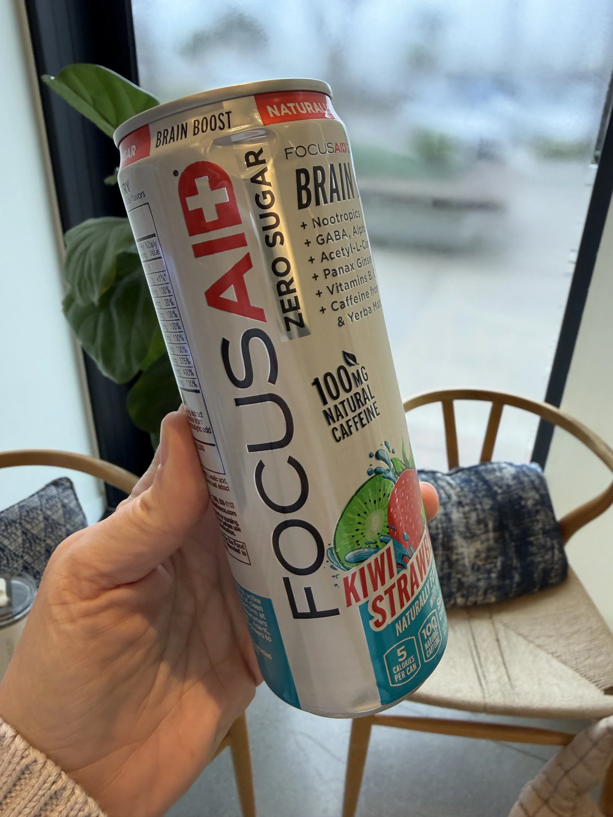A hand holding a can of FocusAid Kiwi Strawberry zero sugar energy drink with a background of chairs, a table, and a large window with a blurred outdoor view.