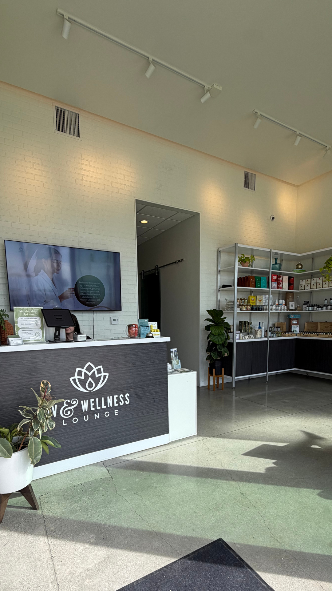 Reception area of a wellness lounge with a black front desk displaying a white logo and the words 'Wellness Lounge'. Shelves with health products and a large flat-screen TV on the wall.