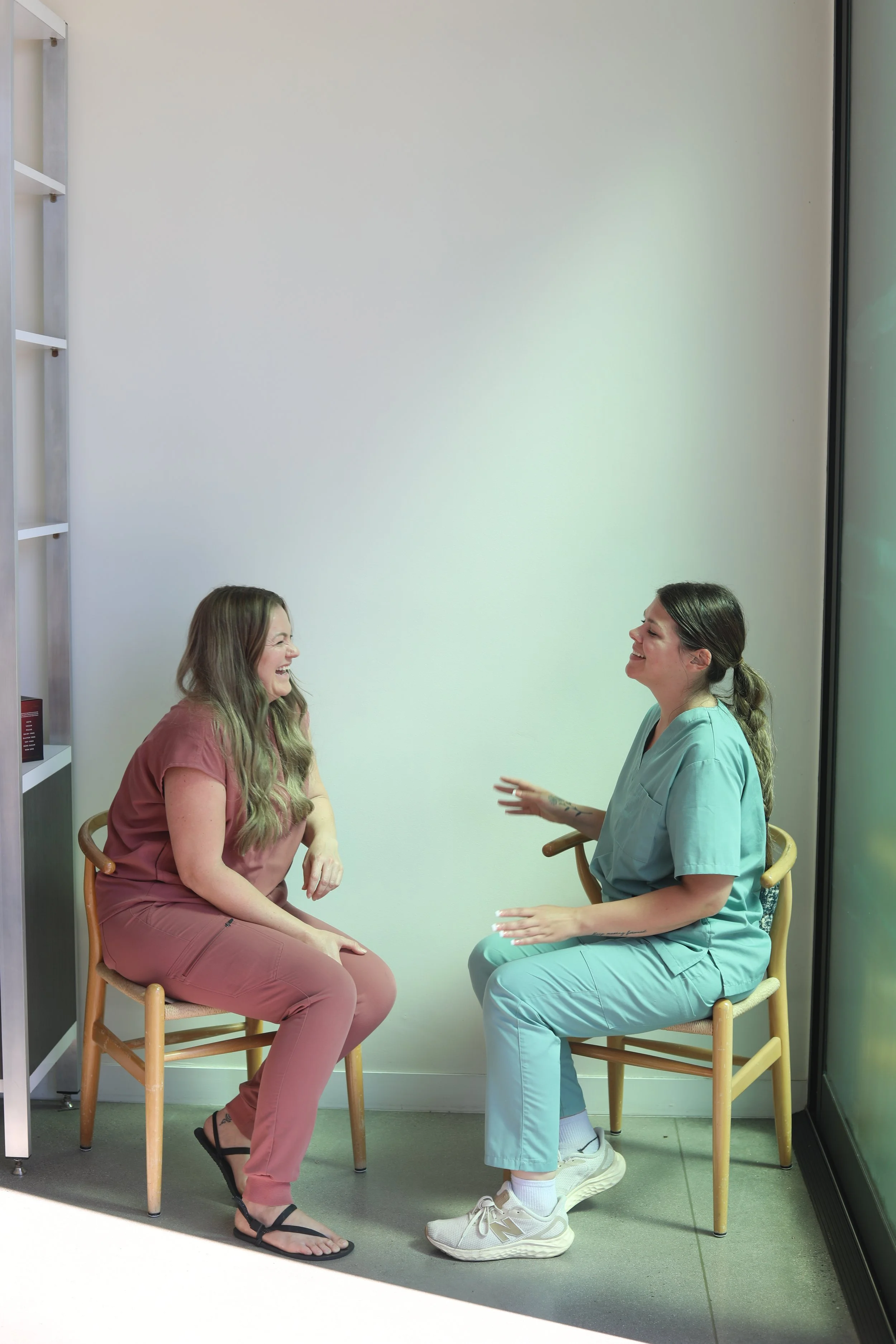 Two women, one in pink scrubs and the other in light blue scrubs, sitting on chairs facing each other, smiling and laughing in a room with white walls and large window.