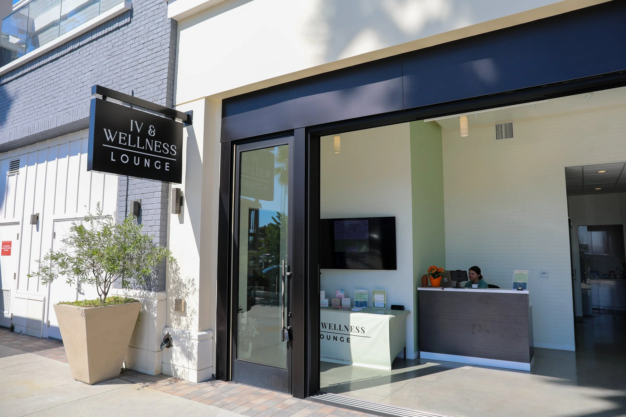 Exterior view of IV & Wellness Lounge entrance with black sign, glass door, white interior, reception desk, and TV inside.