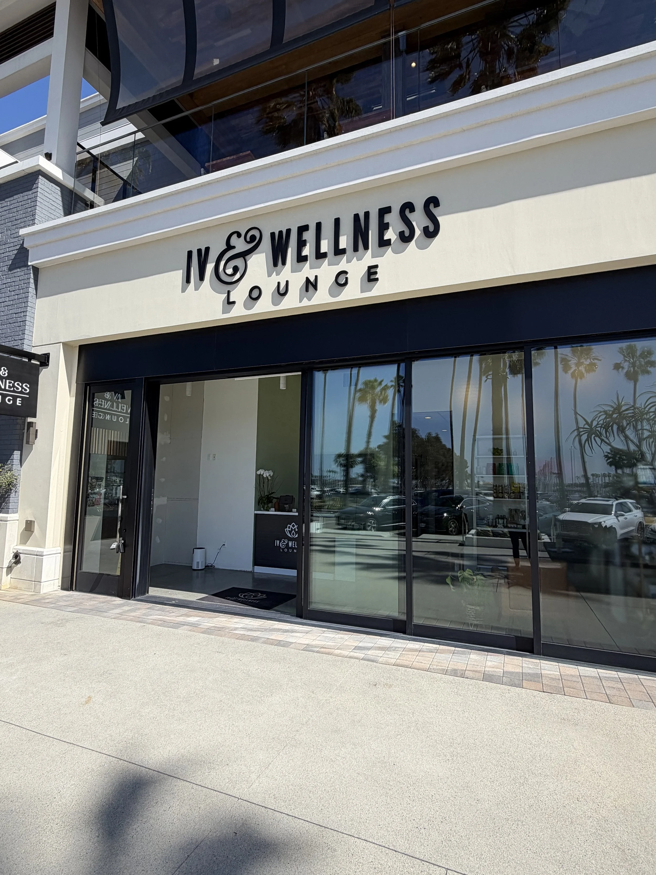 Exterior of IV & Wellness Lounge, a wellness center with large glass windows, palm trees reflections, and a sign above the entrance.