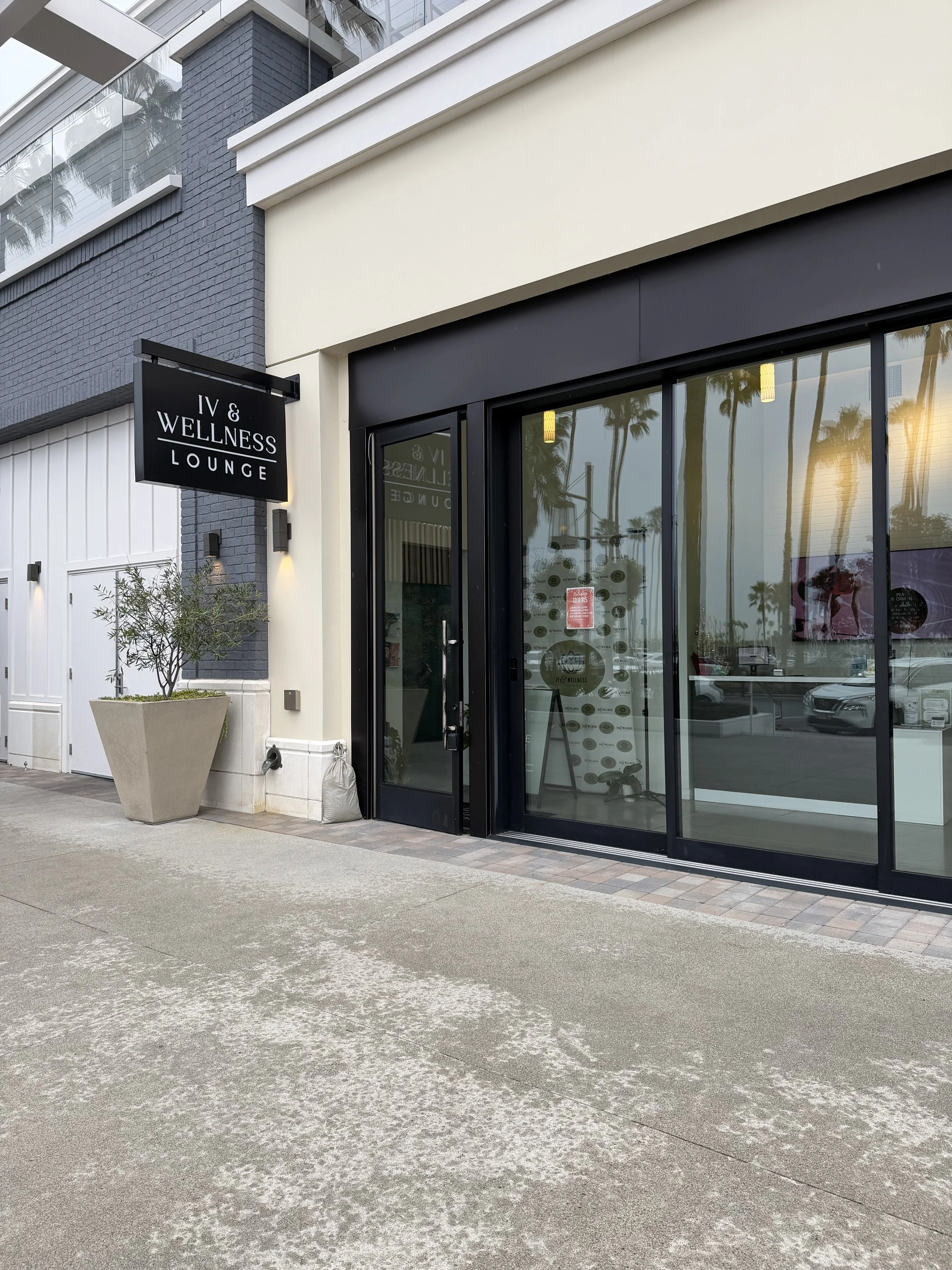 Exterior of a wellness lounge with black sliding glass doors, white walls, a black sign reading 'IV & Wellness Lounge', a potted plant, and palm trees reflected in the glass.